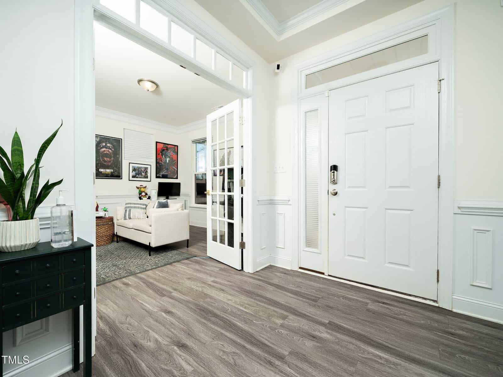A bright entryway with light wood floors, white walls, and a white front door. A small green table with a plant and bottled water sits on the left. Through French doors, a living room with a sofa and wall art is visible.