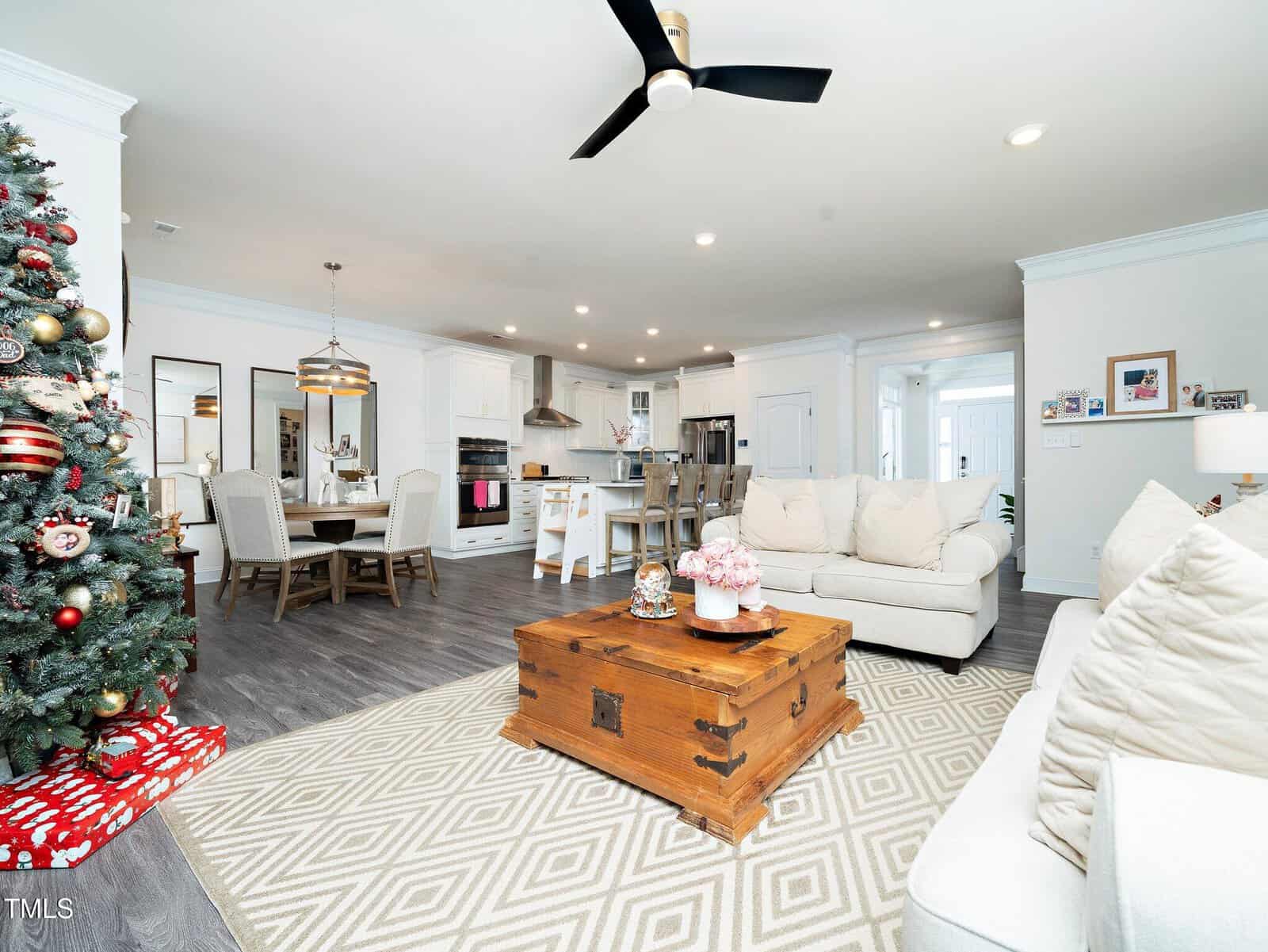 Bright living room and kitchen with white cabinets, beige couches, a wooden coffee table, dining table with chairs, and a decorated Christmas tree with presents beside it. The floor is dark wood and the space is open and modern.