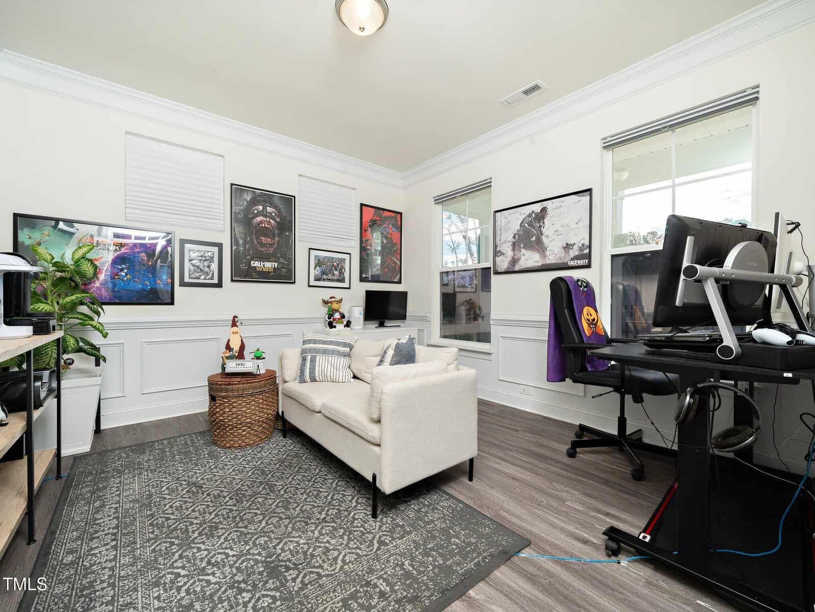 A modern home office with a small beige sofa, desk with computer setup, and multiple gaming and pop culture posters on the walls. Two windows let in natural light. A patterned rug covers part of the wooden floor.