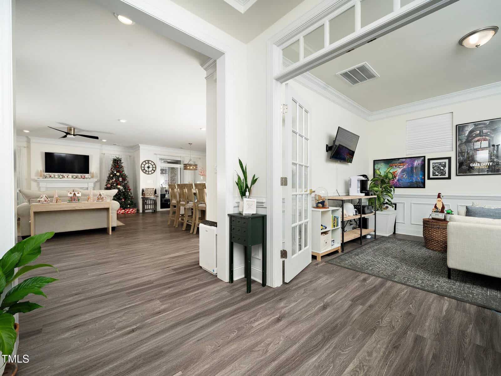 View of an open-plan living space with wood-style flooring, a living room with a Christmas tree and couches, a dining area, and a separate office area with a desk, artwork, and a couch. Several plants are visible.