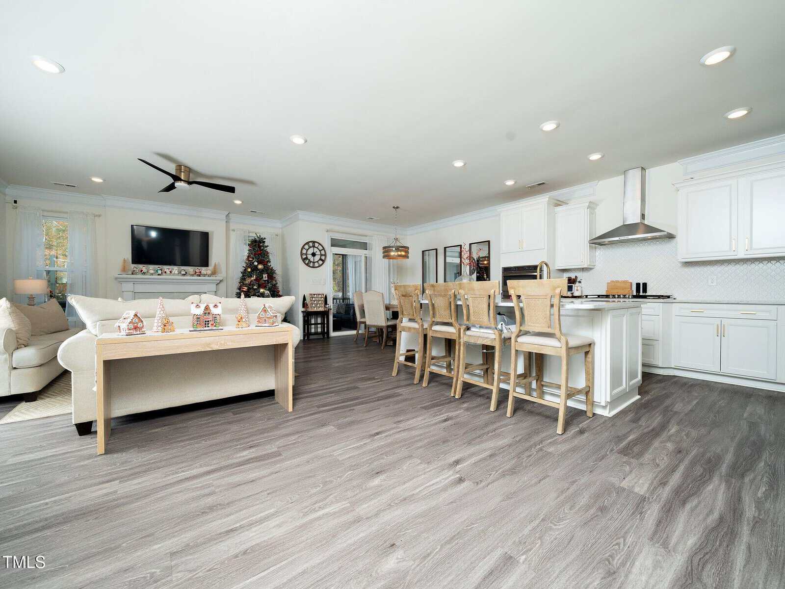 Open-concept living room and kitchen with white cabinetry, light wood flooring, bar stools, a Christmas tree, and holiday decorations on a table. Ceiling fan, TV, and dining area are also visible.