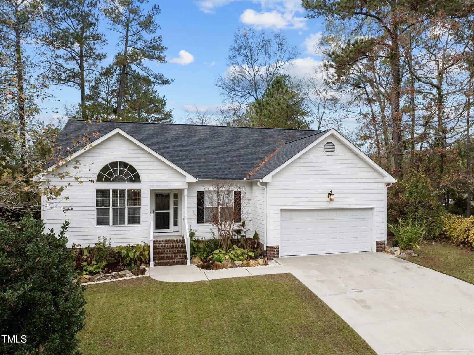 Nestled on 2766 Courtney Court in Creedmoor, this single-story white house with a dark roof features an attached two-car garage. The front yard boasts a small lawn and a few shrubs, while trees surround the property under a partly cloudy sky.