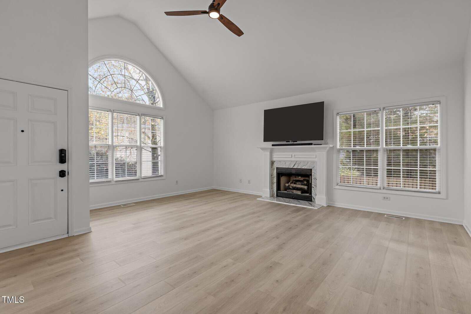 A spacious living room with light wood flooring and white walls. It features a high ceiling with a ceiling fan, a fireplace with a TV above it, large windows with natural light, and a white-paneled door on the left.