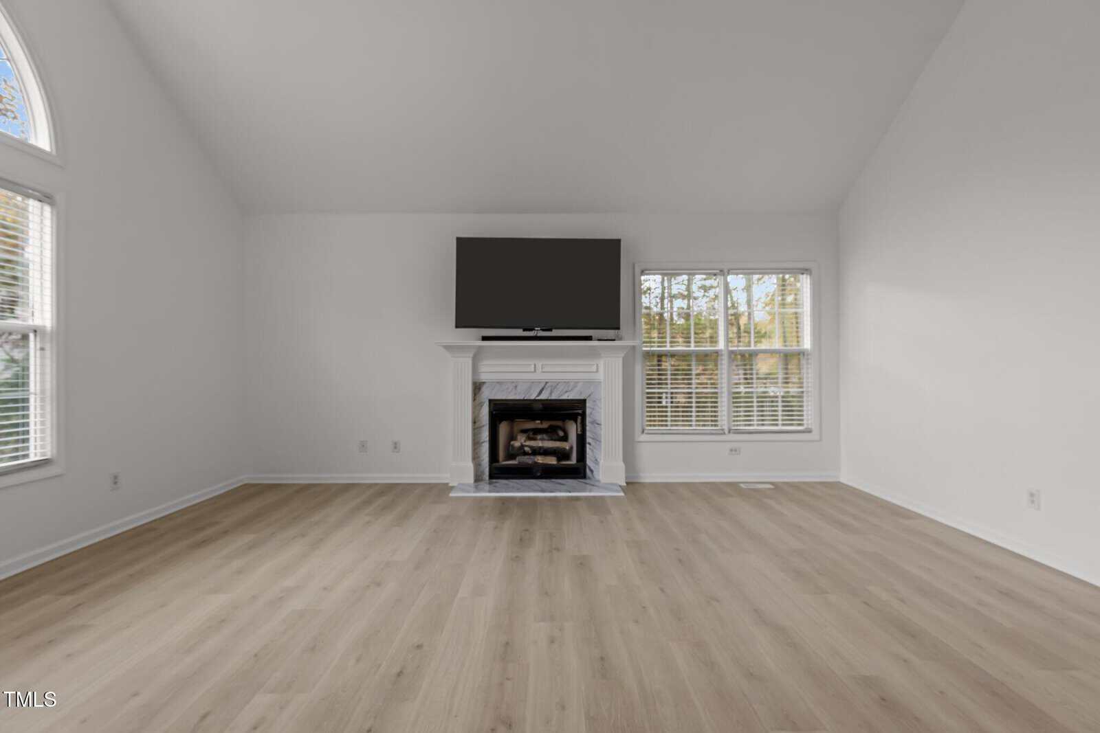 A spacious, empty living room with light wooden floors and white walls. A large flat-screen TV is mounted above a fireplace. Two large windows on each side let in natural light.