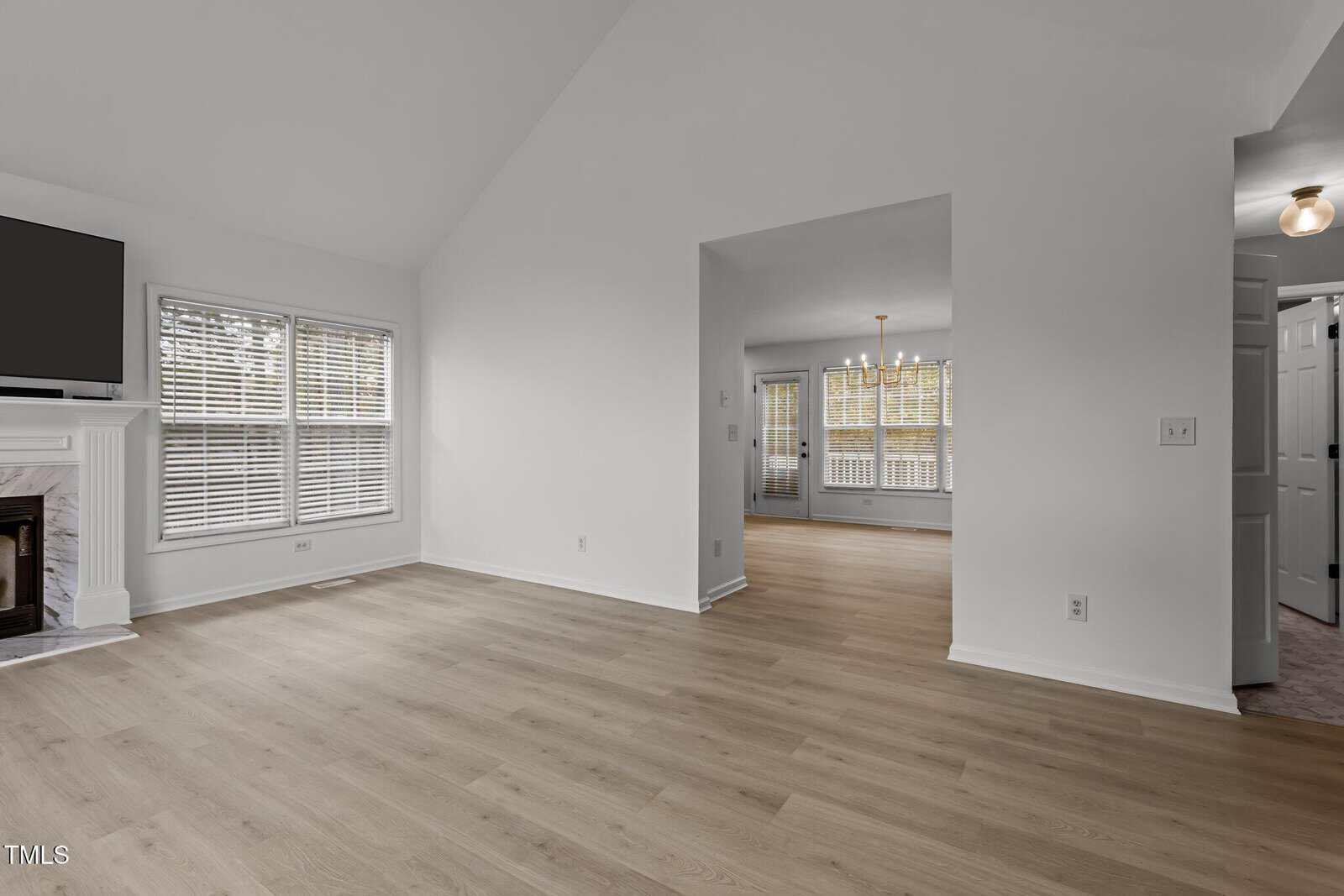 Spacious, well-lit living room with light wood flooring, white walls, and large windows. A fireplace is on the left, and an open doorway leads to another room with a light fixture.