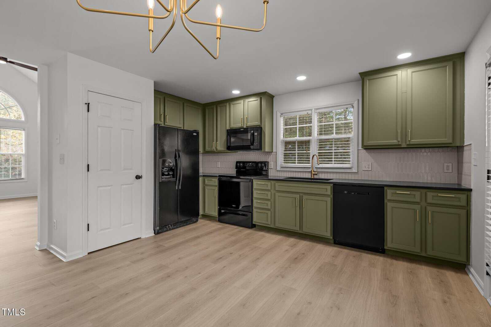 A modern kitchen with green cabinets, black appliances, and light wood flooring. A white door is on the left, and a window above the sink offers natural light. A gold chandelier hangs from the ceiling.