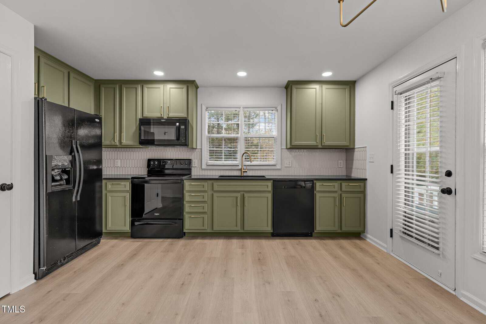 A modern kitchen with olive green cabinets, black appliances, and a wooden floor. There is a large window above the sink, a glass door to the right, and recessed lighting illuminating the room.
