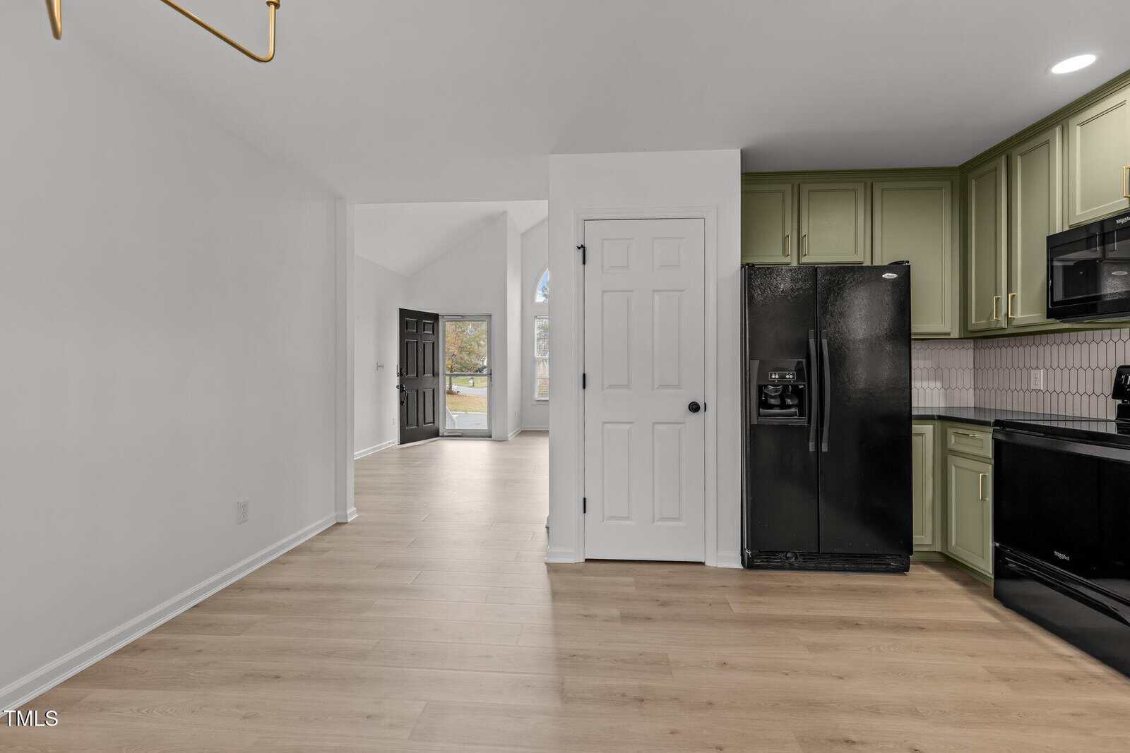 View of a modern kitchen with green cabinets, black appliances, and light wood flooring. A white door leads to another room, visible through an open doorway showing a black door and arched window. The space is bright and airy.