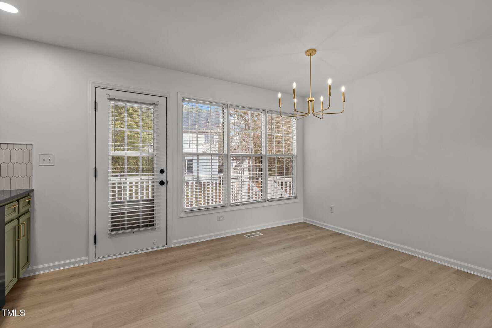 Bright, empty dining room with large windows and a modern gold chandelier. Hardwood floors and a glass door leading outside. White walls and green cabinets visible on the left.