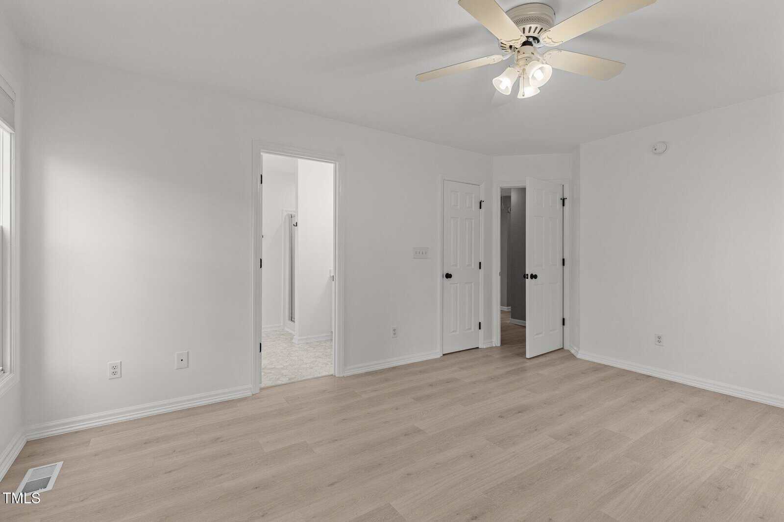A bright, empty room with light wood flooring and white walls. The ceiling has a white fan with lights. Doors lead to a bathroom and another room. Natural light comes from a window on the left.