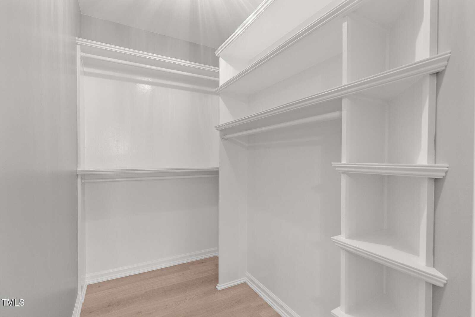A spacious, white walk-in closet with multiple shelves and hanging rods. The closet has a light wooden floor and offers ample storage space for clothing and accessories.