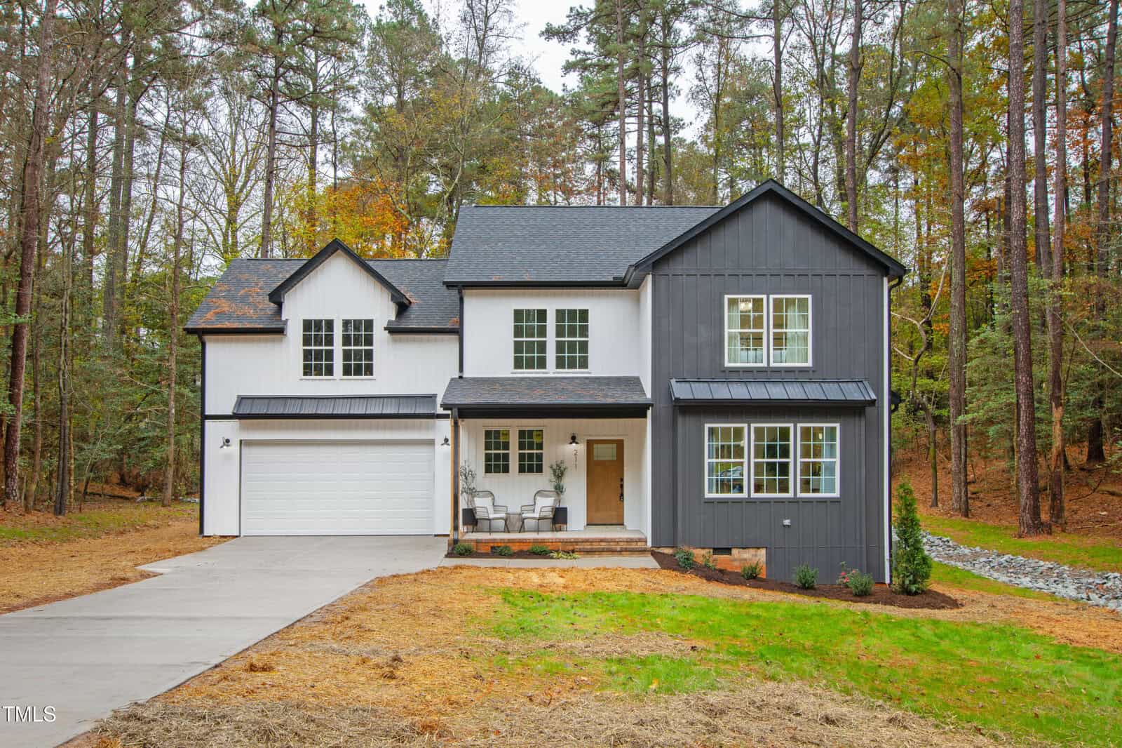 Two-story modern house with white and dark gray siding, a double garage, and a front porch with two chairs. The house is surrounded by trees and has a grass lawn with a concrete driveway.