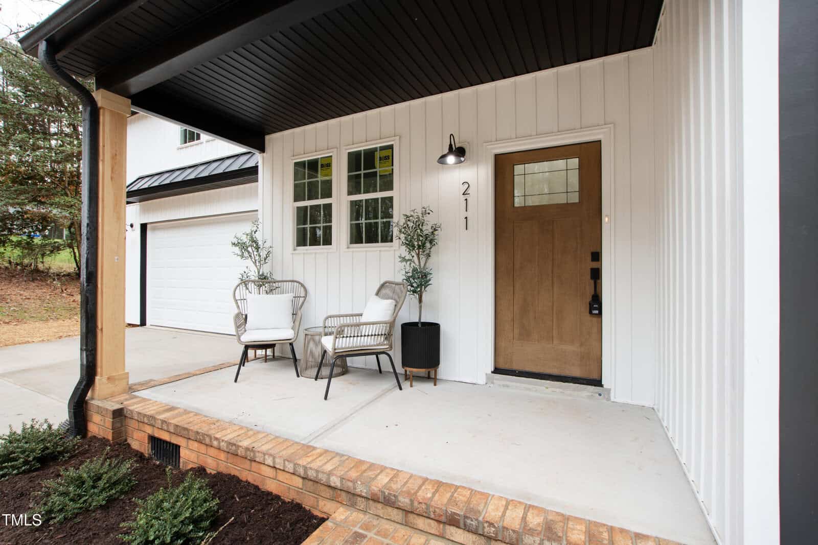 A modern house front porch with white vertical siding, a wooden door with a window, two chairs, a small table, potted plants, a garage door on the left, and a light above the house number 211.