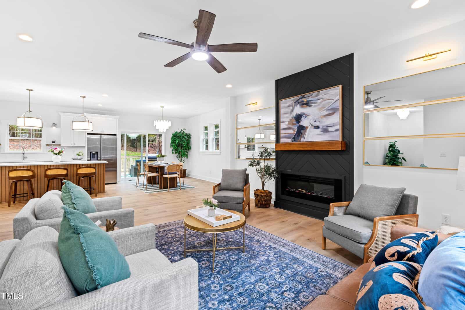 A modern open-concept living room and kitchen with light wood floors, a blue patterned rug, grey and brown furniture, a black accent fireplace, and a kitchen island with three stools. Large windows provide natural light.