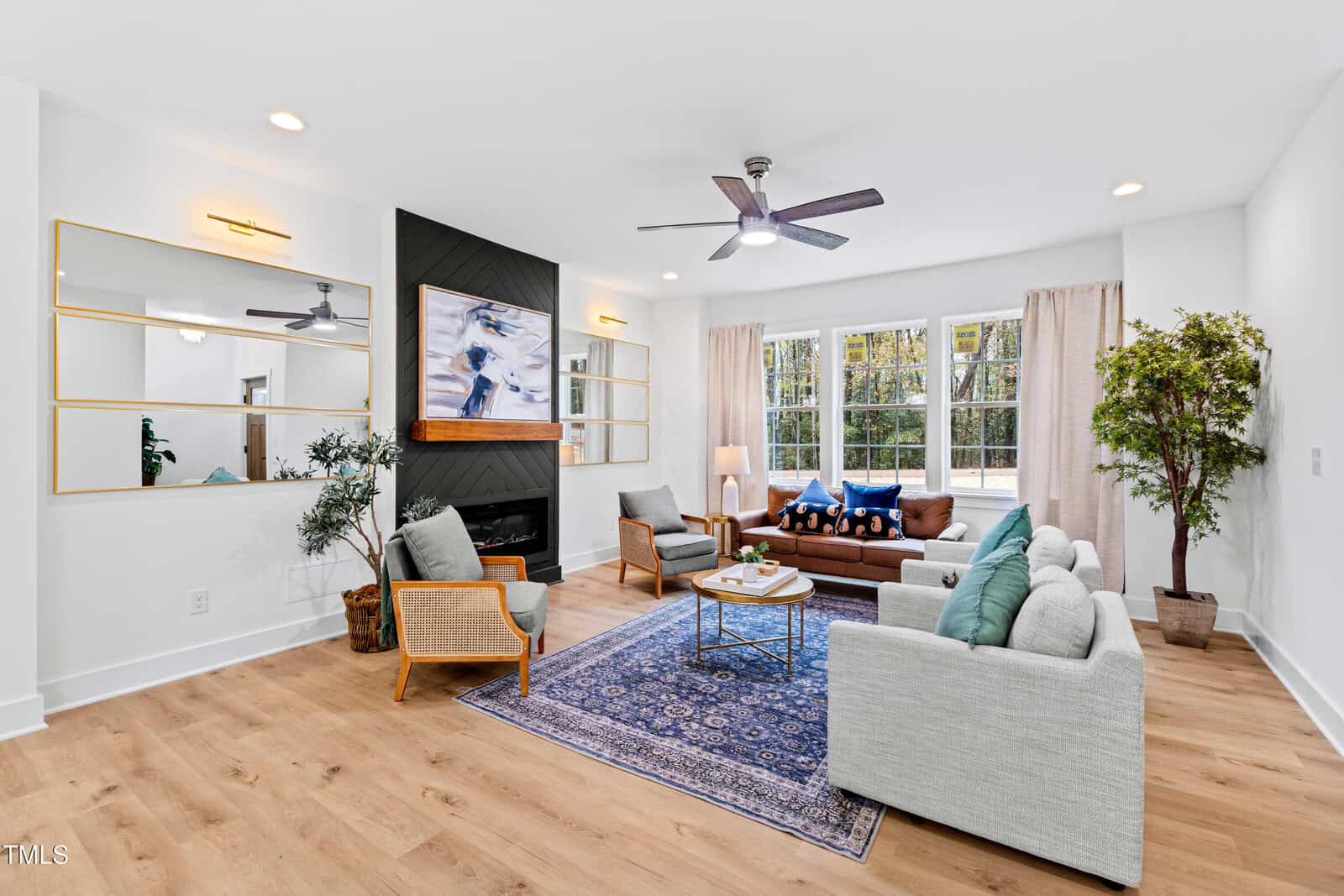A bright, modern living room with light wood floors, a gray sofa, two armchairs, a blue area rug, a brown couch, a coffee table, large windows with curtains, a fireplace, and mirrors on the wall.