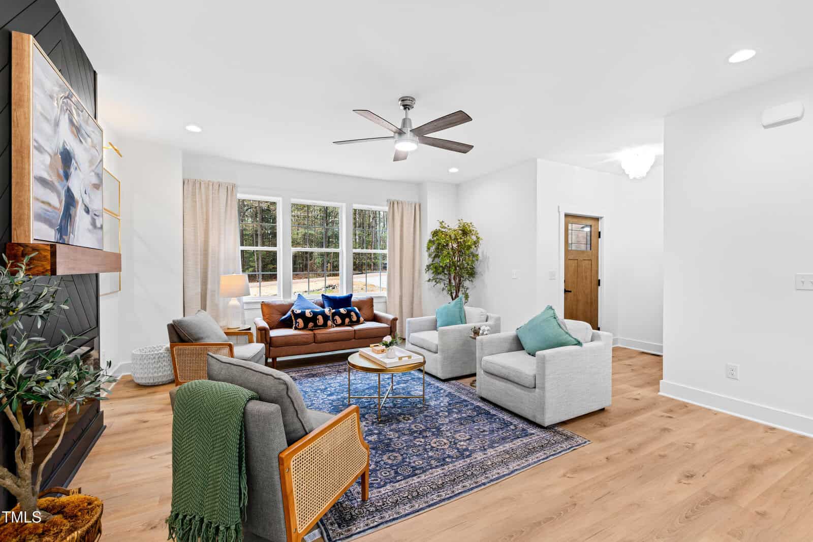A modern living room with a brown leather sofa, two gray armchairs, a blue rug, a wooden coffee table, large windows with beige curtains, a ceiling fan, and wood flooring. A plant and decor items are also visible.