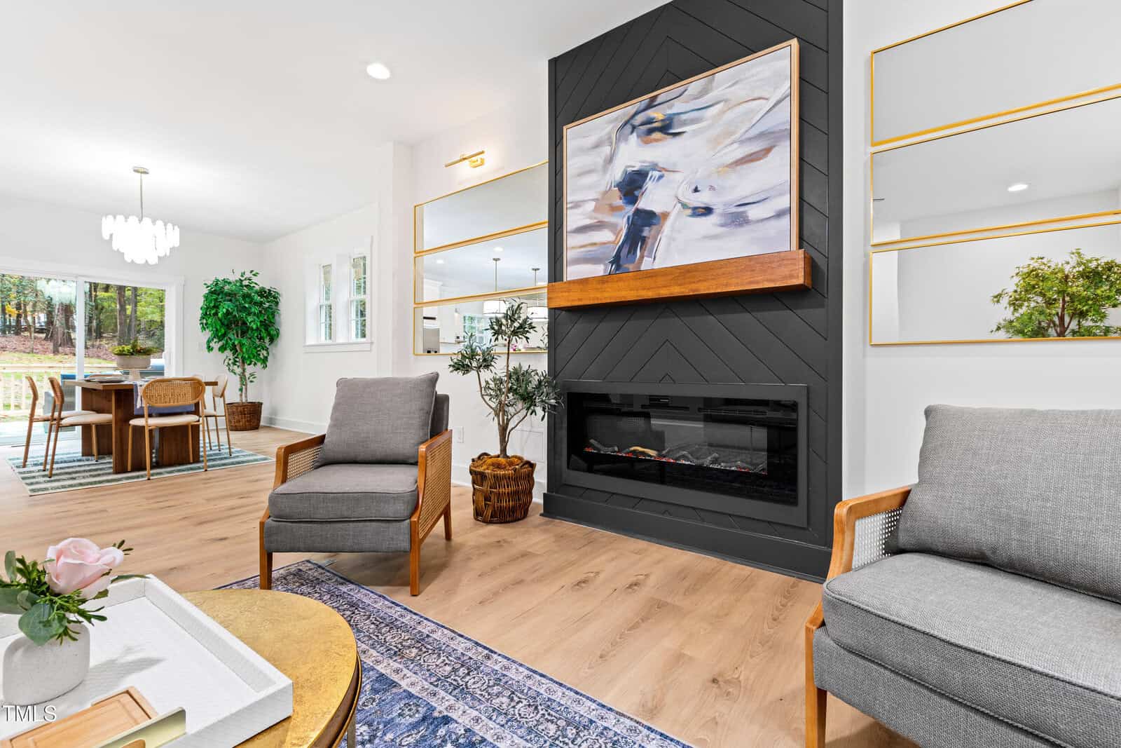 Modern living room with a dark accent fireplace, abstract art, gray chairs, and wood floors. Large mirrors and bright windows reflect light. Dining area and greenery visible in background. Cozy, contemporary décor.