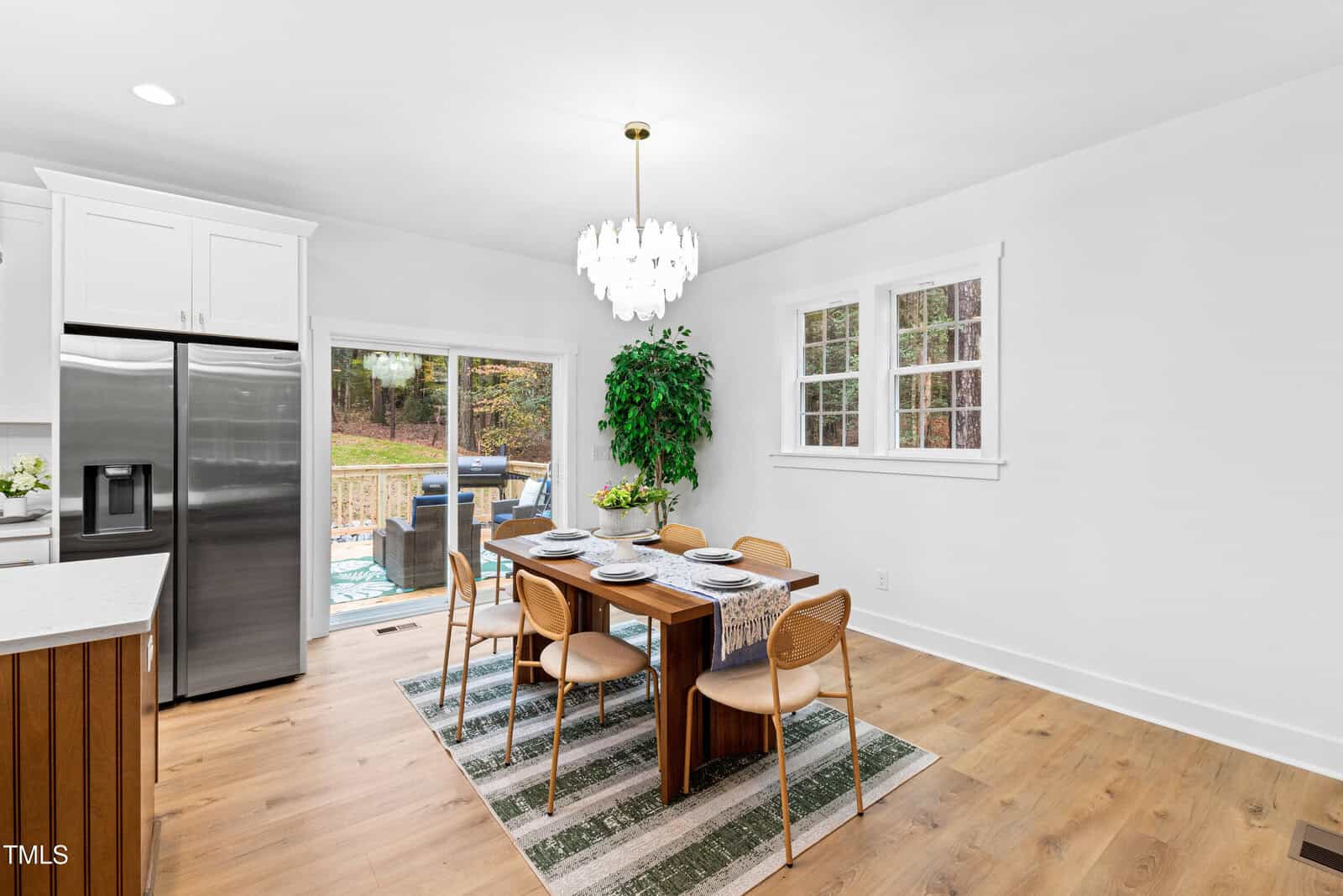Modern dining area with wooden table set for six, light wood chairs, a striped rug, large green plant, stainless fridge, white cabinets, chandelier, and glass doors opening to a patio with grill and outdoor seating.