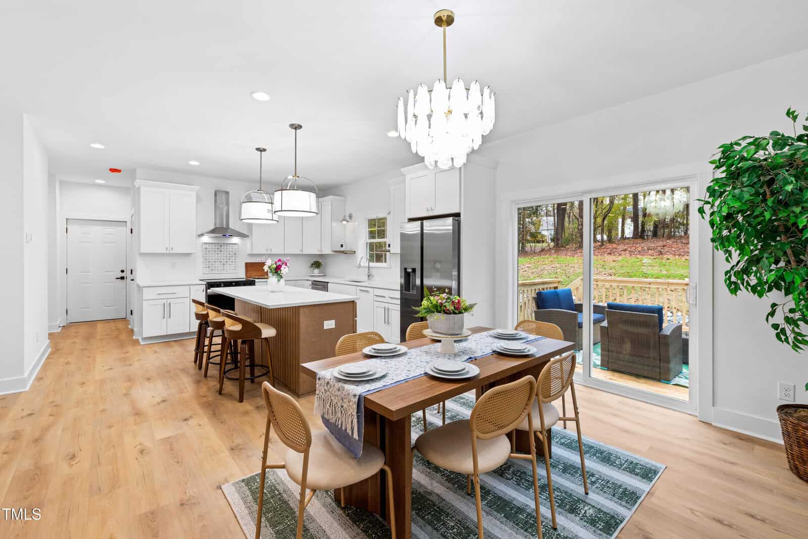 Bright modern kitchen and dining area with wood floors, white cabinets, an island with stools, a set dining table, a chandelier, sliding doors to a patio, and a large window with a view of the backyard.