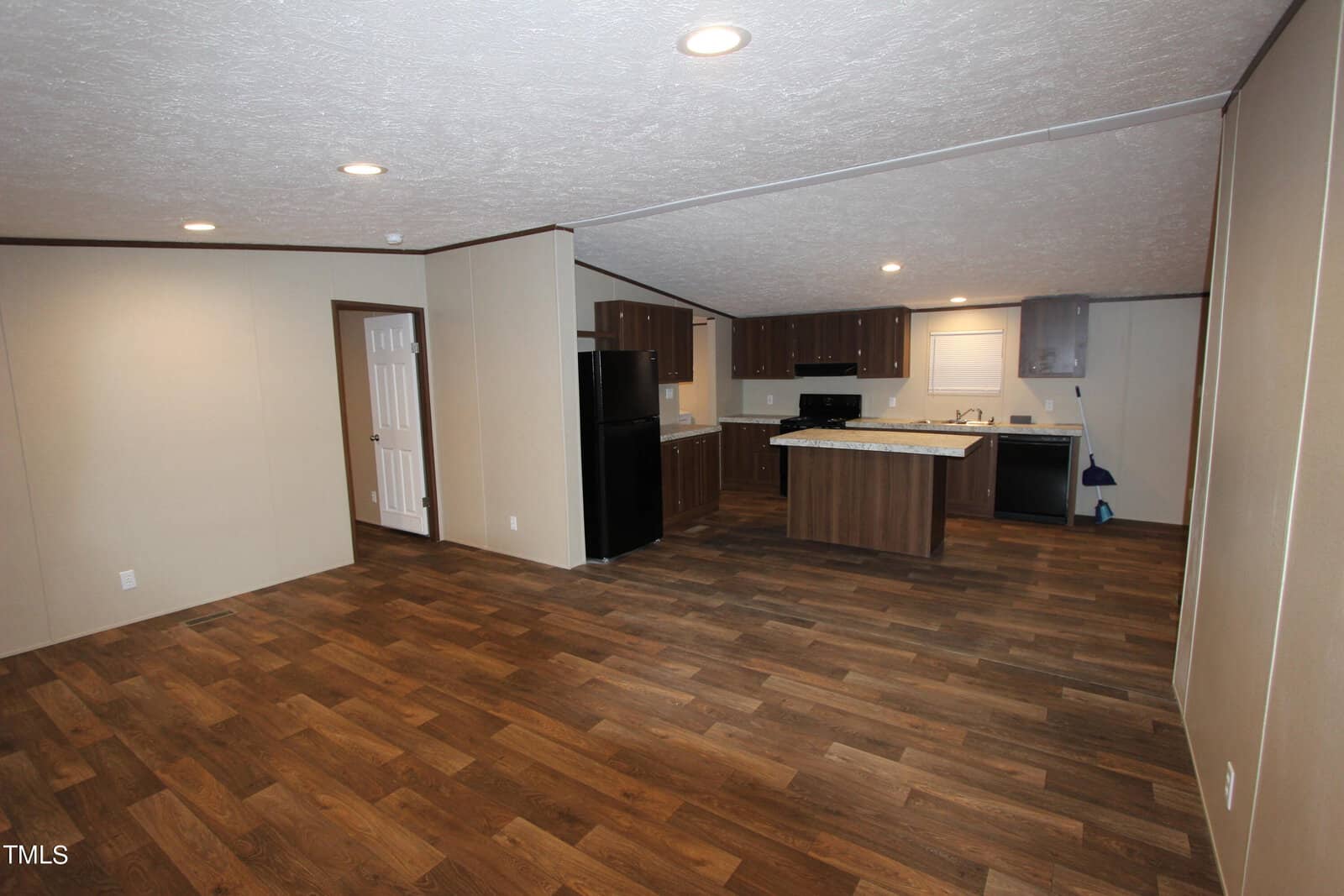 Spacious open-plan living area with wood flooring, modern kitchen featuring dark cabinetry, a central island, black appliances, and recessed ceiling lights. Beige walls and a white door in the background complete the setting.