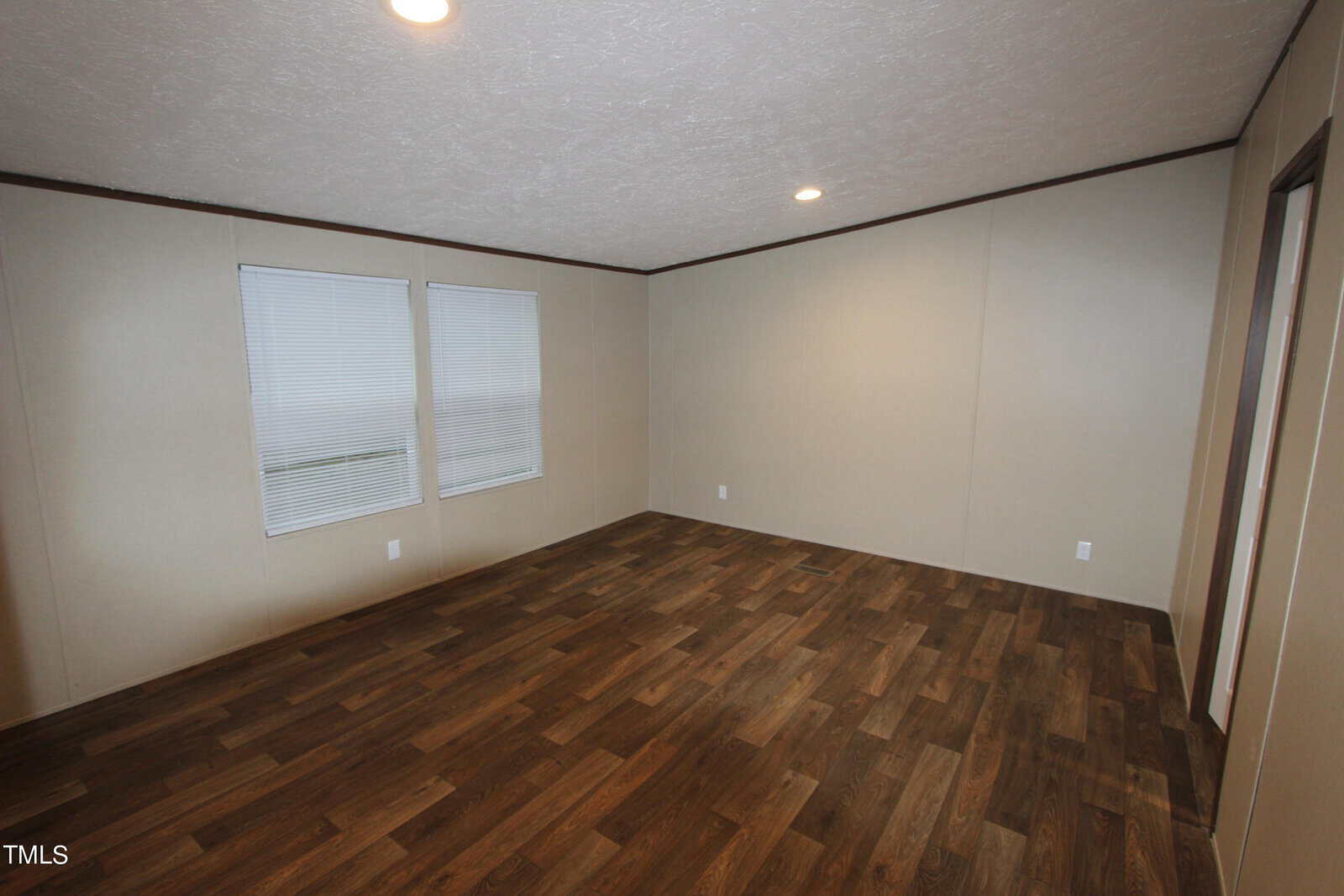 An empty room with beige walls and a dark wood floor. Two windows with blinds are on one wall, and ceiling lights are turned on, creating a bright, clear atmosphere.