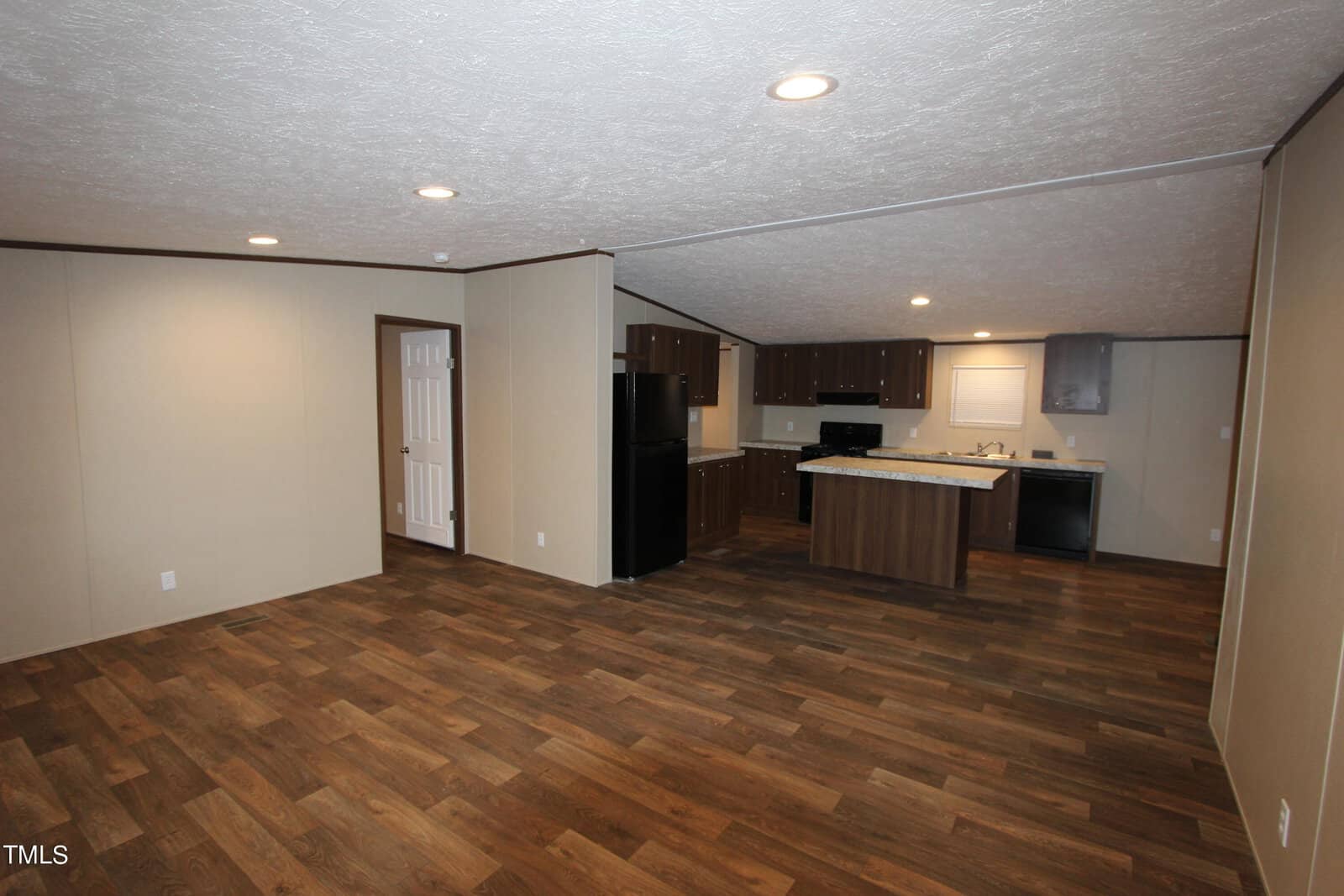 A spacious open-plan living area with wooden flooring, beige walls, and ceiling lights. It features a kitchen at the back with dark wood cabinets, a fridge, oven, and dishwasher. A white door leads to another room.