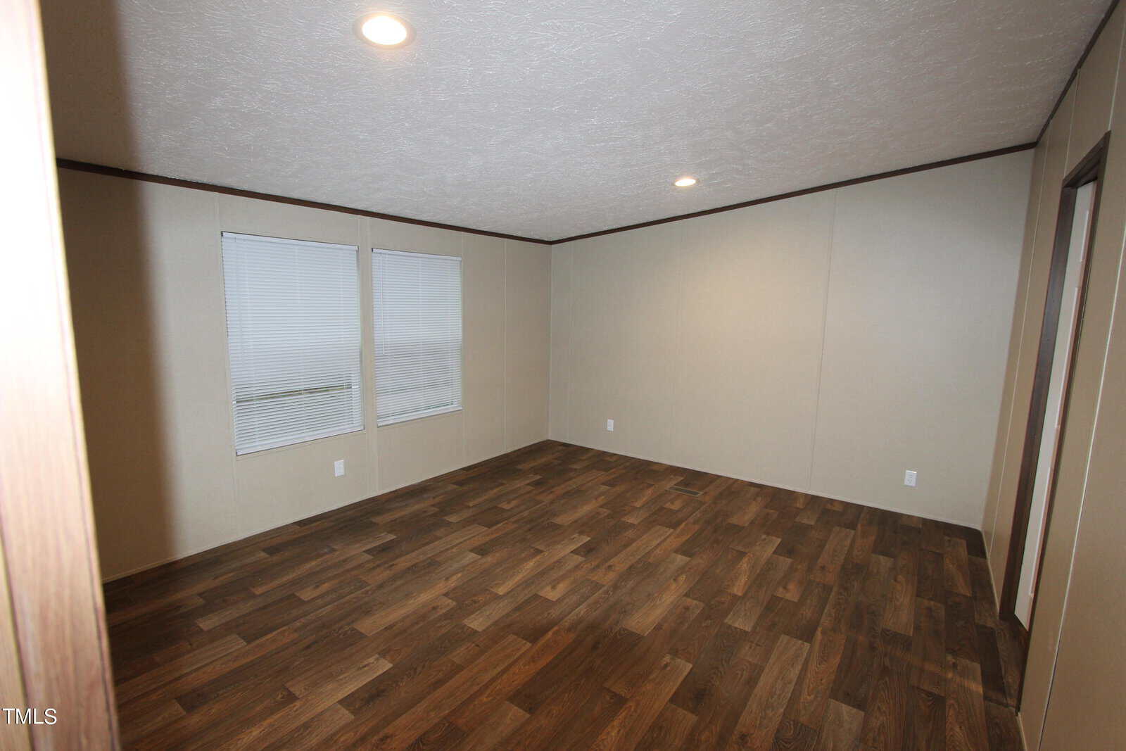 An empty room with beige walls and wooden flooring. Two windows are on one wall, partially covered by blinds. The ceiling features recessed lighting.