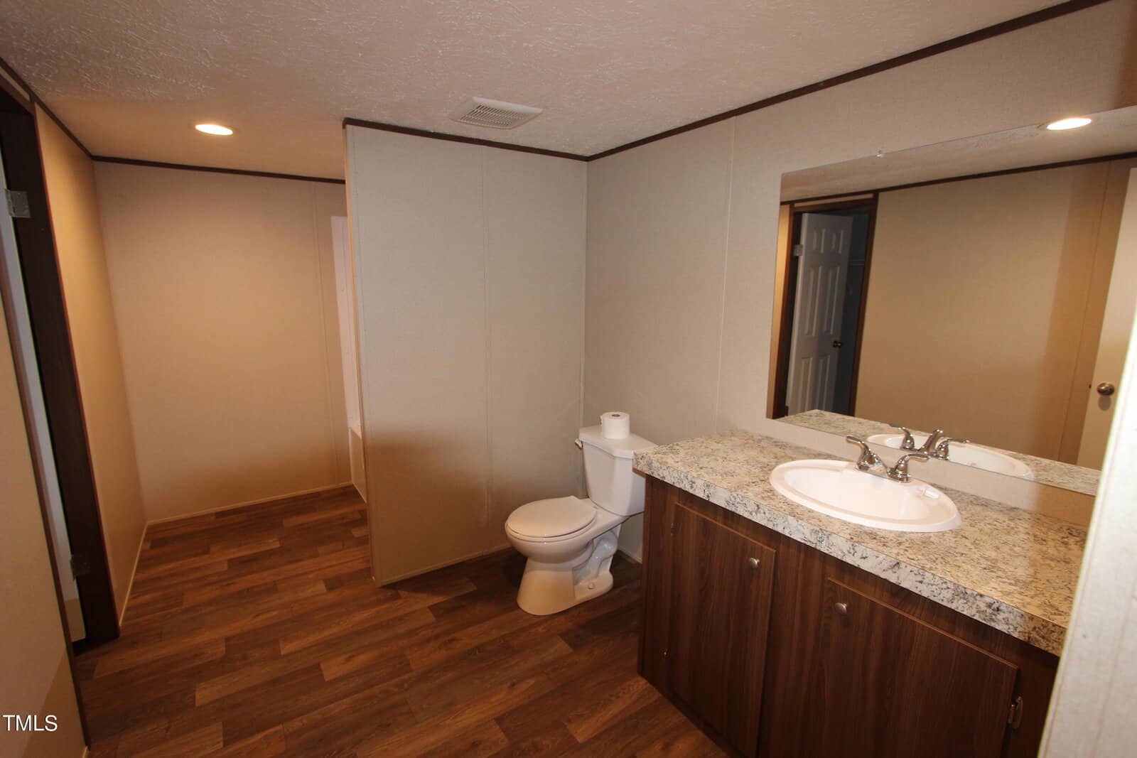 A bathroom with beige walls and wood flooring. There's a white toilet next to a granite-topped vanity with a sink and faucet. A large mirror is mounted above the sink, and recessed lighting is installed in the ceiling.