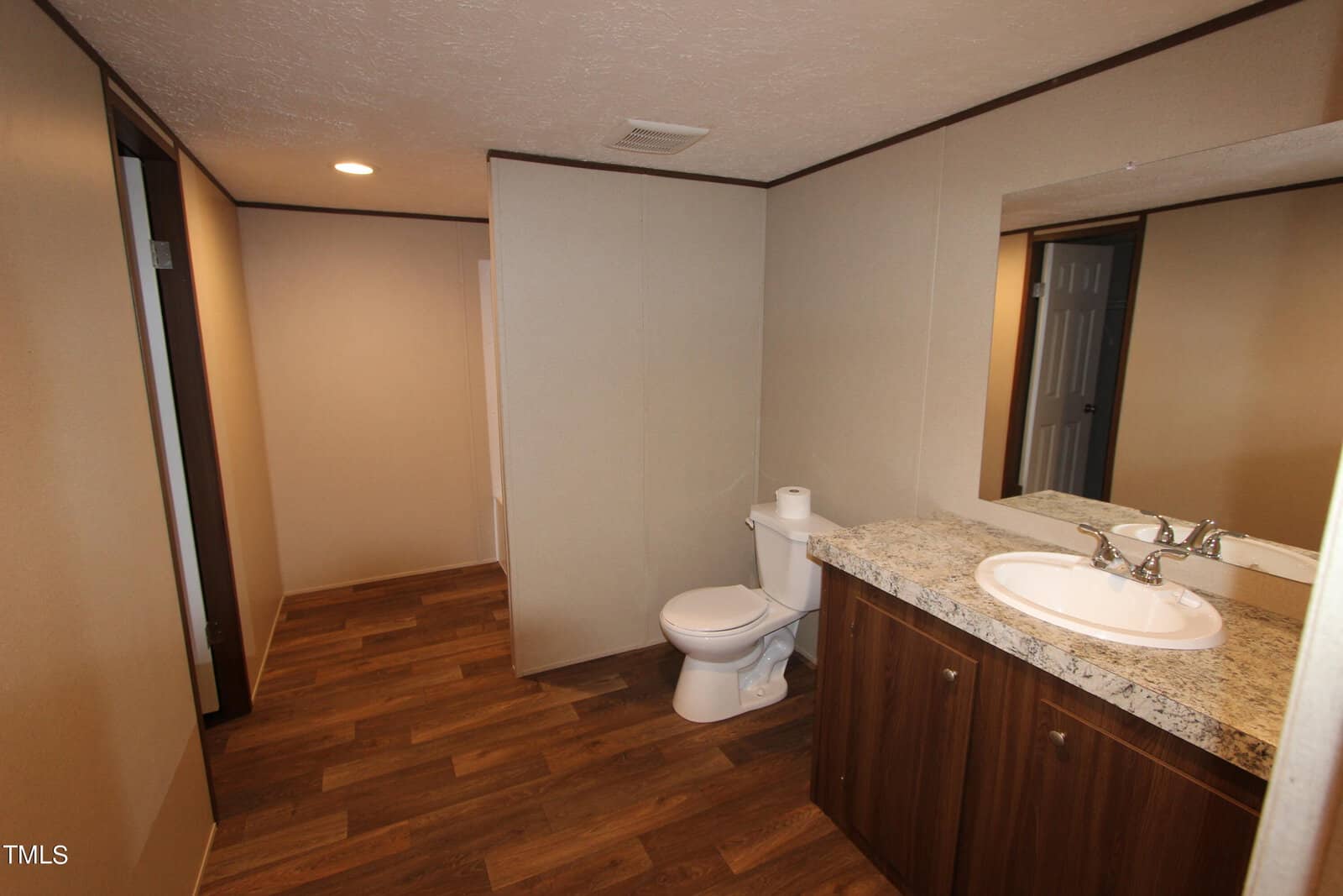 A modern bathroom with wooden flooring, featuring a white toilet and granite-topped vanity with a sink and faucet. The walls are beige, and there is a large mirror above the vanity. Recessed lighting illuminates the room.
