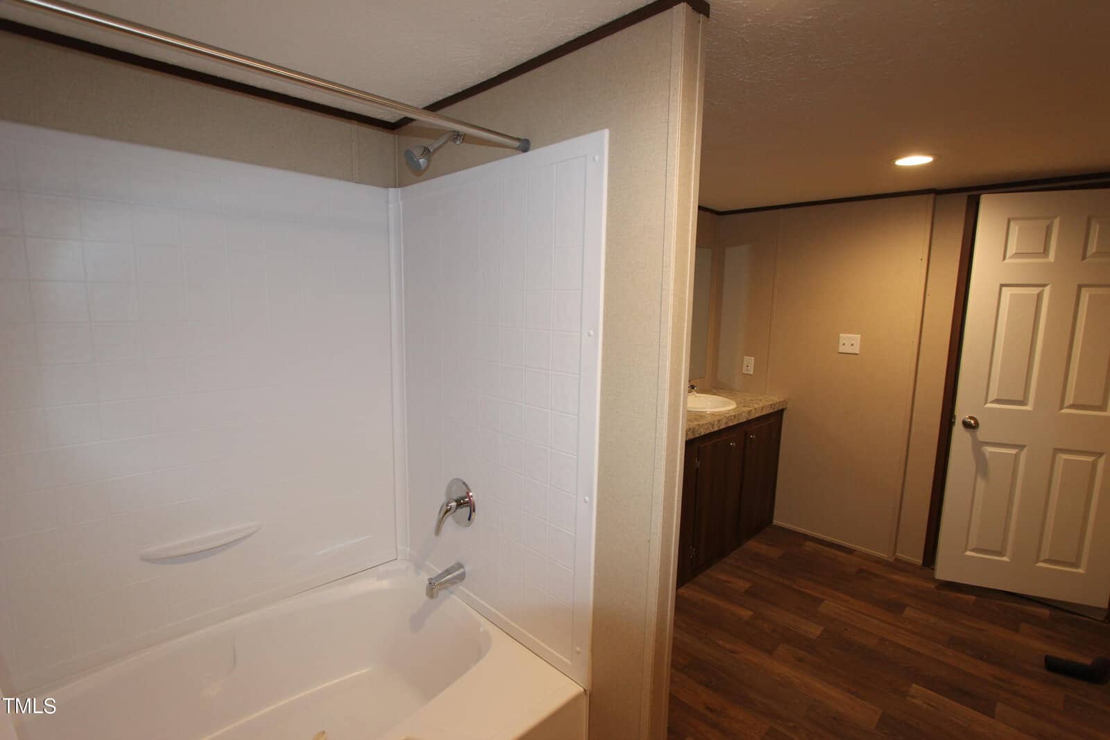 A bathroom with a white bathtub and shower combo on the left, featuring a beige wall. The floor is wood-textured, and the room leads to a white door and a double-sink vanity with a mirror on the right.