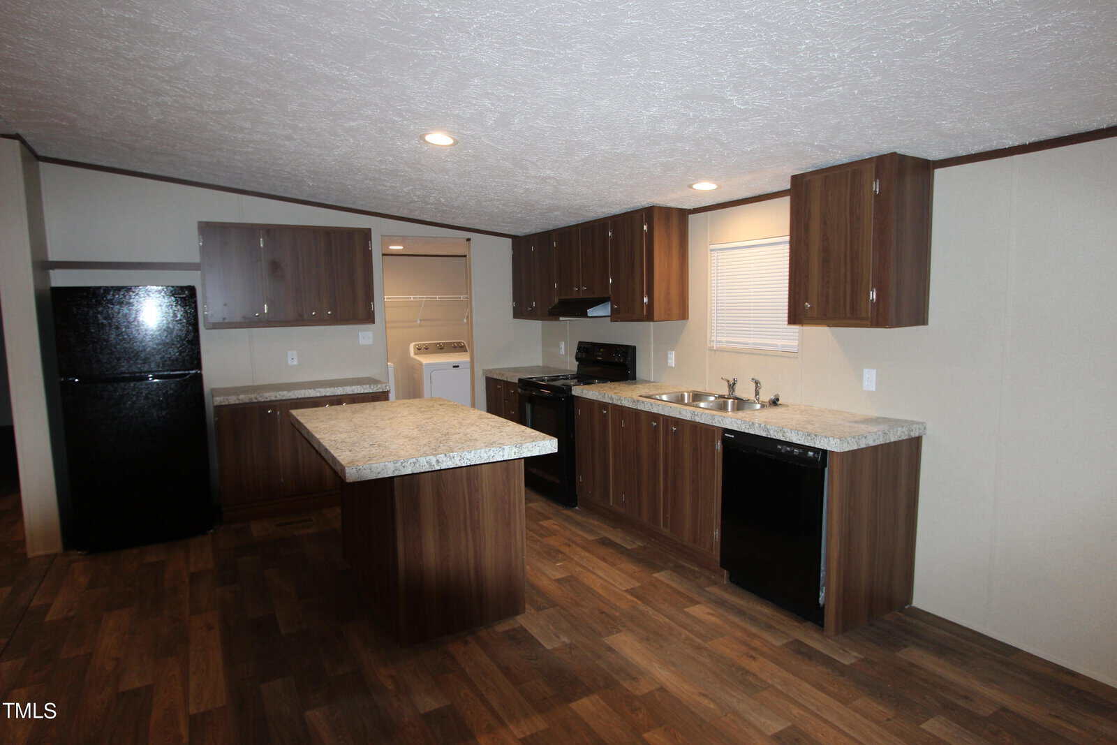 Modern kitchen with dark wood cabinets, marble countertops, and stainless steel appliances. Features an island, black fridge, dishwasher, stove, and a view into a laundry room. Wood laminate flooring and recessed lighting add warmth and brightness.