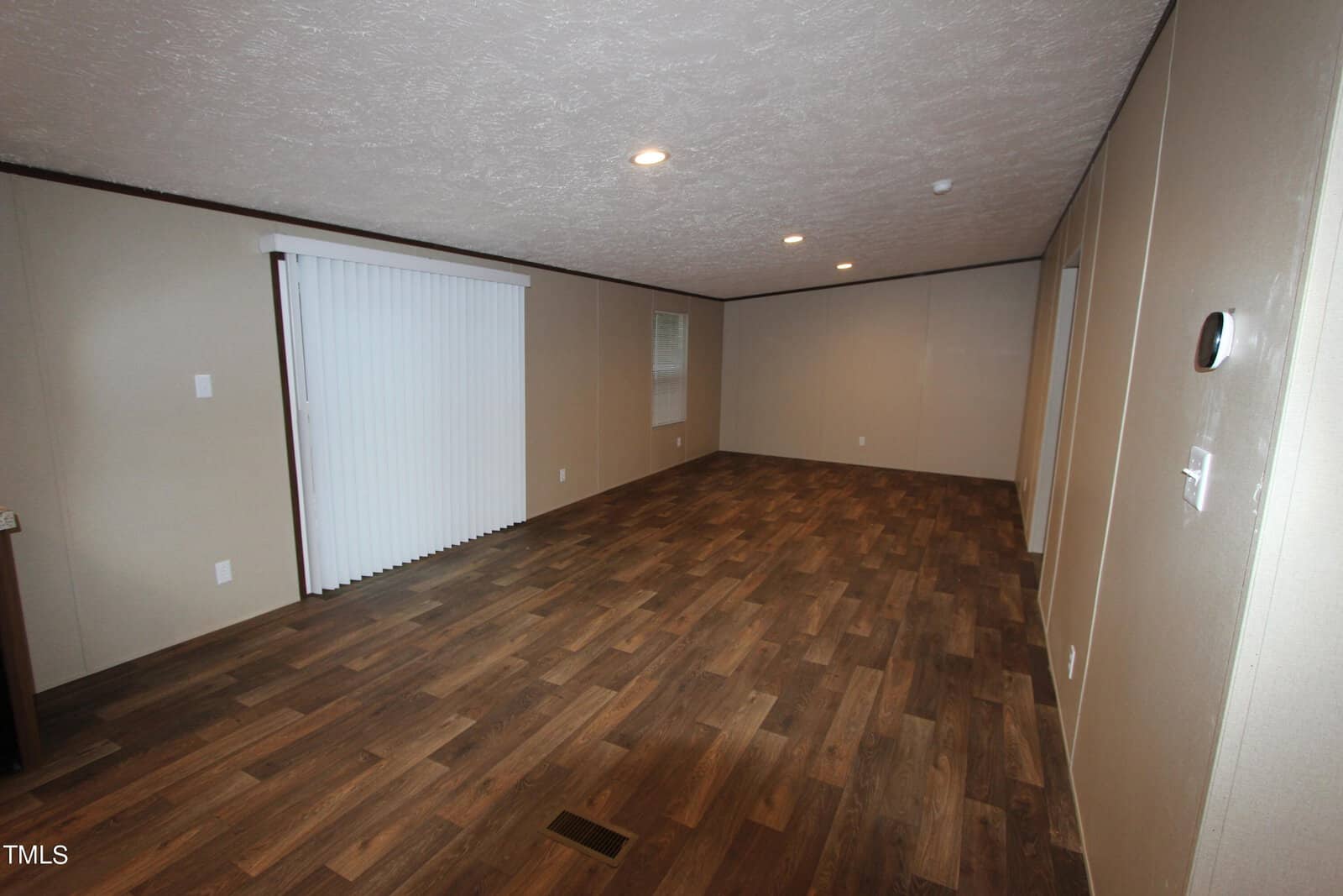 Empty room with wood-patterned flooring and light brown walls. The ceiling is white with a single recessed light. A large window with closed vertical blinds is on one side, and a small wall vent is visible.