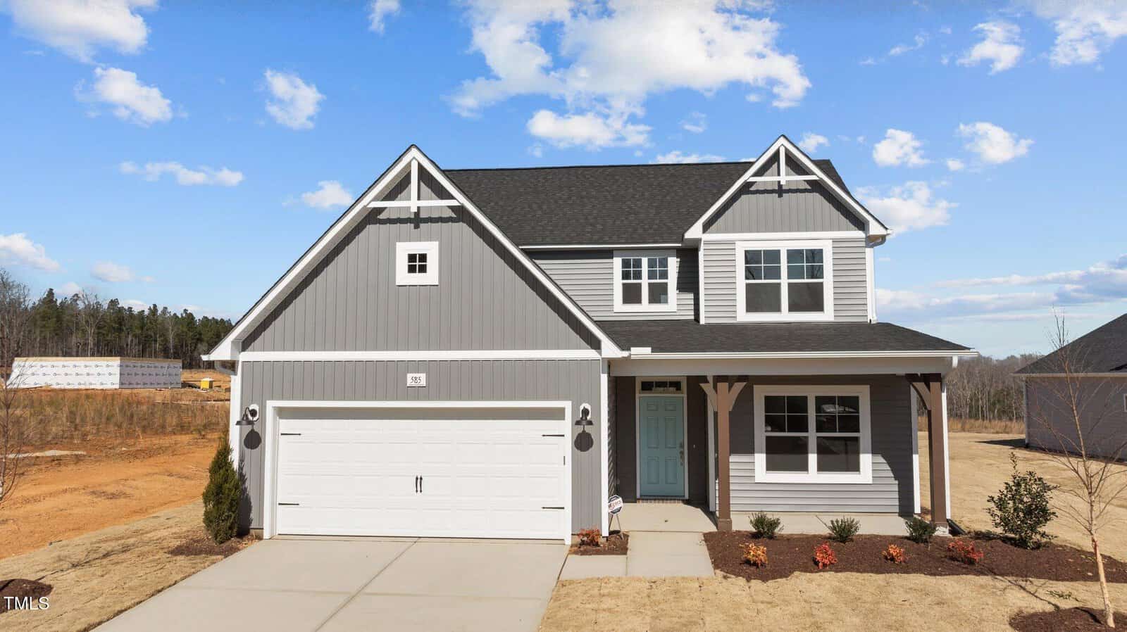 A two-story gray house with white trim, a black roof, and a double garage. There is a small covered porch, minimal landscaping, and a driveway. The sky is partly cloudy, and the lawn is mostly dry grass.