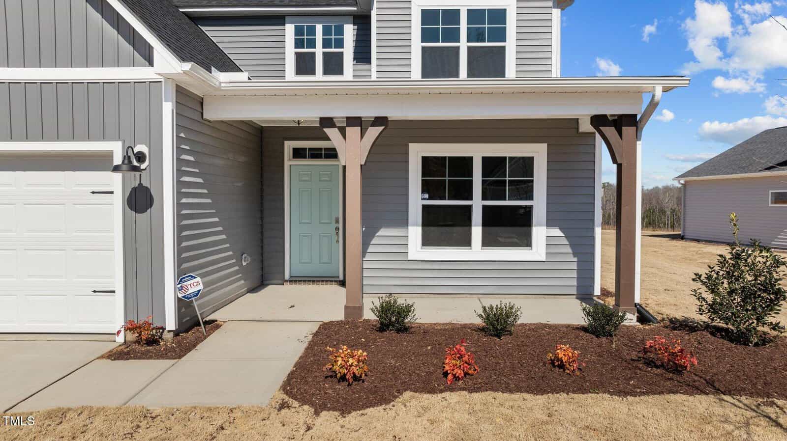 A two-story gray house with white trim, a teal front door, covered porch, and a landscaped front yard with small shrubs and mulch. A security sign is placed near the garage. The sky is clear and sunny.