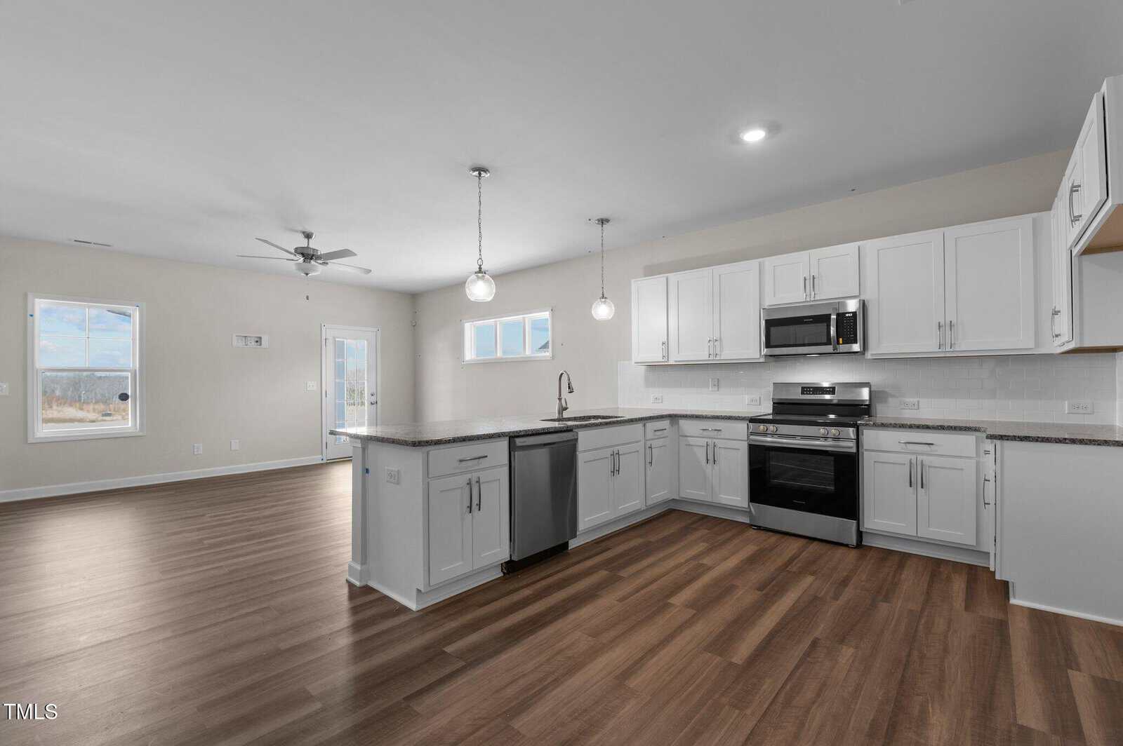 Modern kitchen with white cabinets, stainless steel appliances, granite countertops, and wood flooring, open to a spacious living area with ceiling fan and large windows letting in natural light.