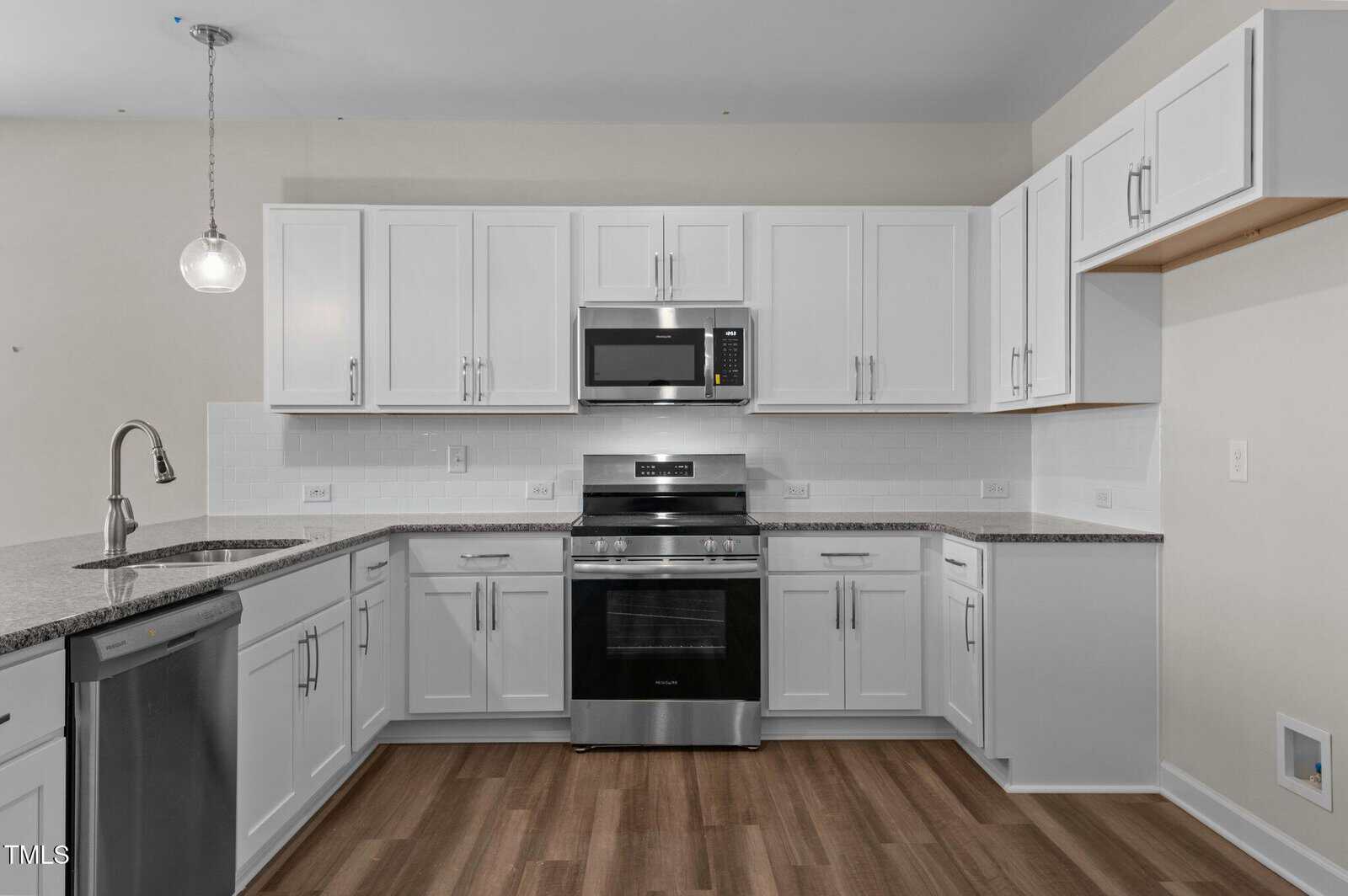 Modern kitchen with white cabinets, stainless steel appliances including a stove, microwave, and dishwasher, gray countertops, tile backsplash, pendant light, and wood-look flooring.