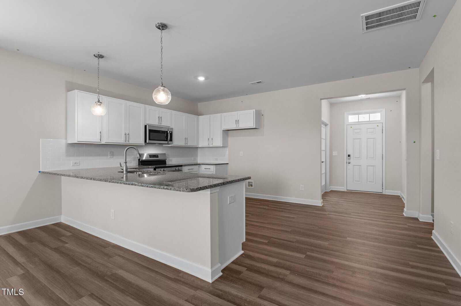 A modern kitchen with white cabinets, stainless steel appliances, and granite countertops. The space features two pendant lights, wood-look flooring, and an open floor plan leading to a front door entryway.