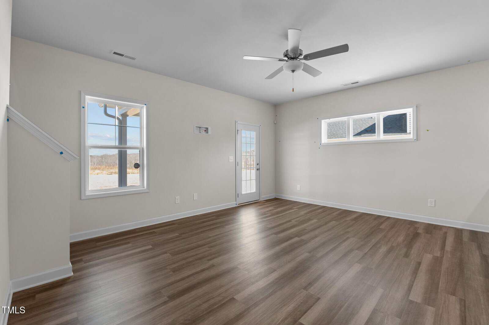 A bright, empty room with light beige walls, wood flooring, three windows, a ceiling fan, and a glass door leading outside. Natural light fills the space.