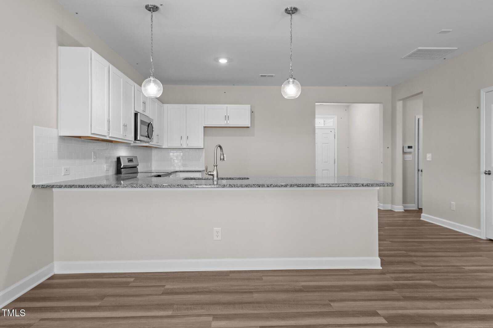 Modern kitchen with white cabinets, stainless steel appliances, granite countertops, subway tile backsplash, pendant lighting, and wood-style flooring. Open layout with neutral walls and visible hallway entrance in the background.