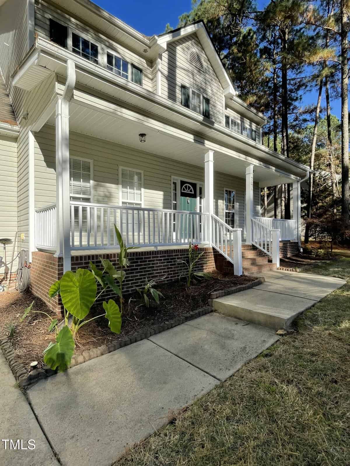 A two-story beige house with white trim and a large covered front porch, white railings, brick steps, and a landscaped garden bed with green plants along the walkway. Tall trees stand in the background.