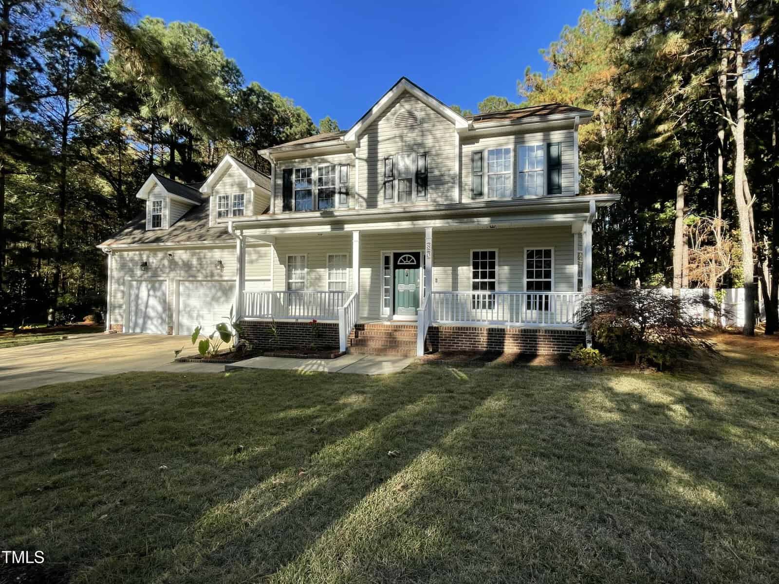A two-story house with light-colored siding, green shutters, a front porch with white railings, and a double garage, surrounded by trees and a well-maintained lawn under a clear blue sky.
