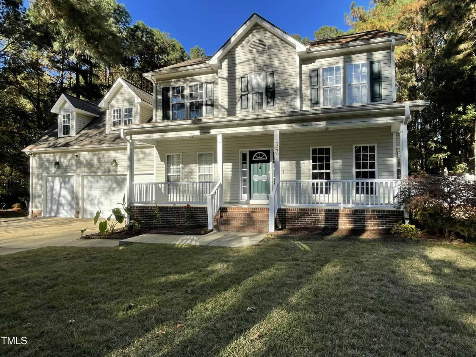 A two-story suburban house with white siding, green shutters, a covered front porch, and a two-car garage, surrounded by trees and a well-kept lawn on a sunny day.