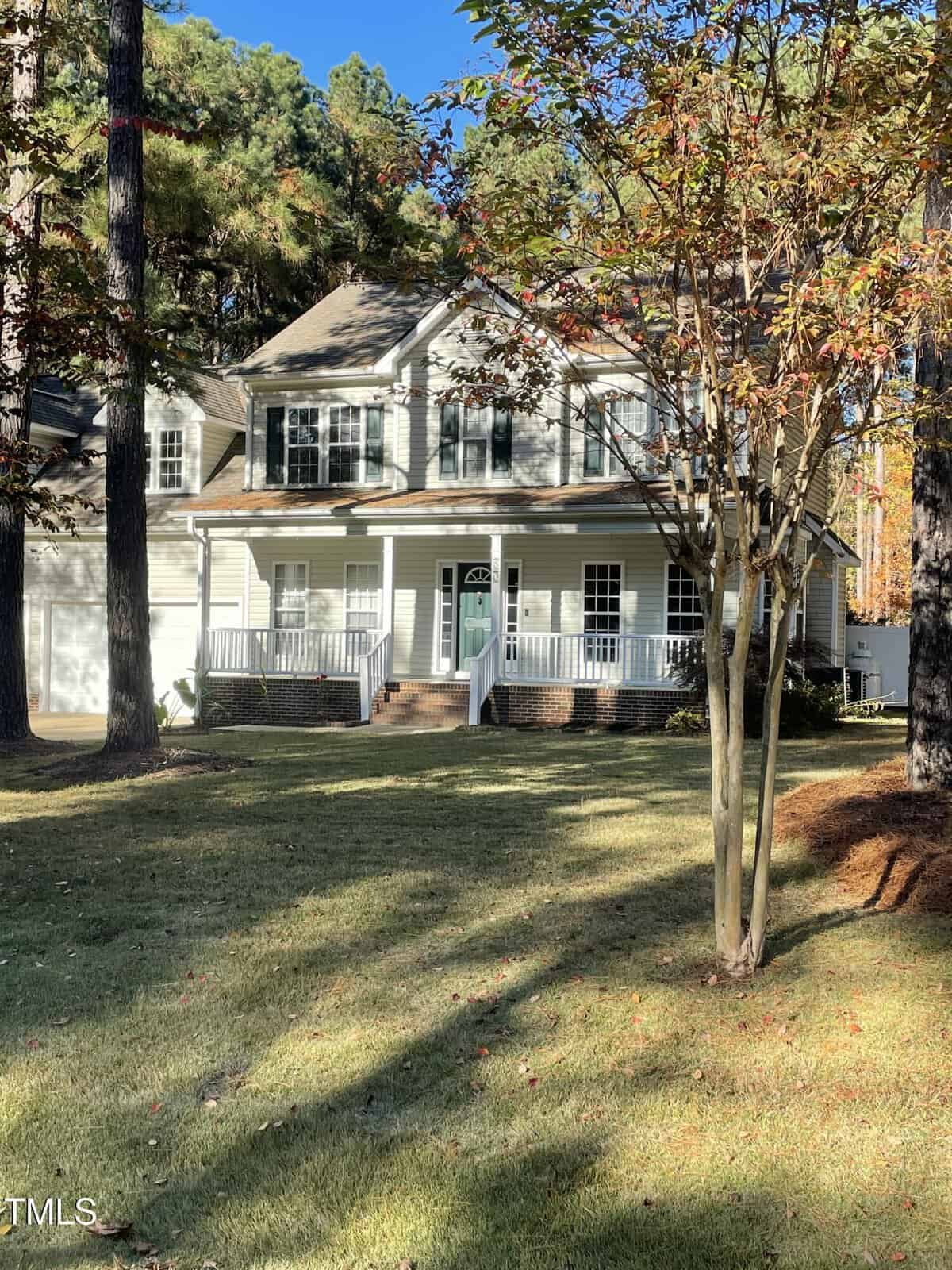 Two-story suburban house with white siding, dark shutters, and a covered front porch, surrounded by trees and a well-kept lawn on a sunny day.