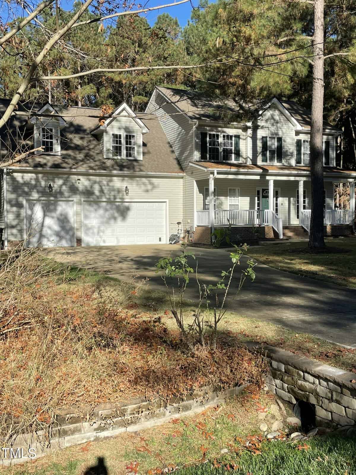Two-story beige house with white trim, a large porch, and three-car garage, surrounded by tall pine trees. Sunlight casts shadows on the driveway and grassy yard, with some autumn leaves on the ground and bushes in front.