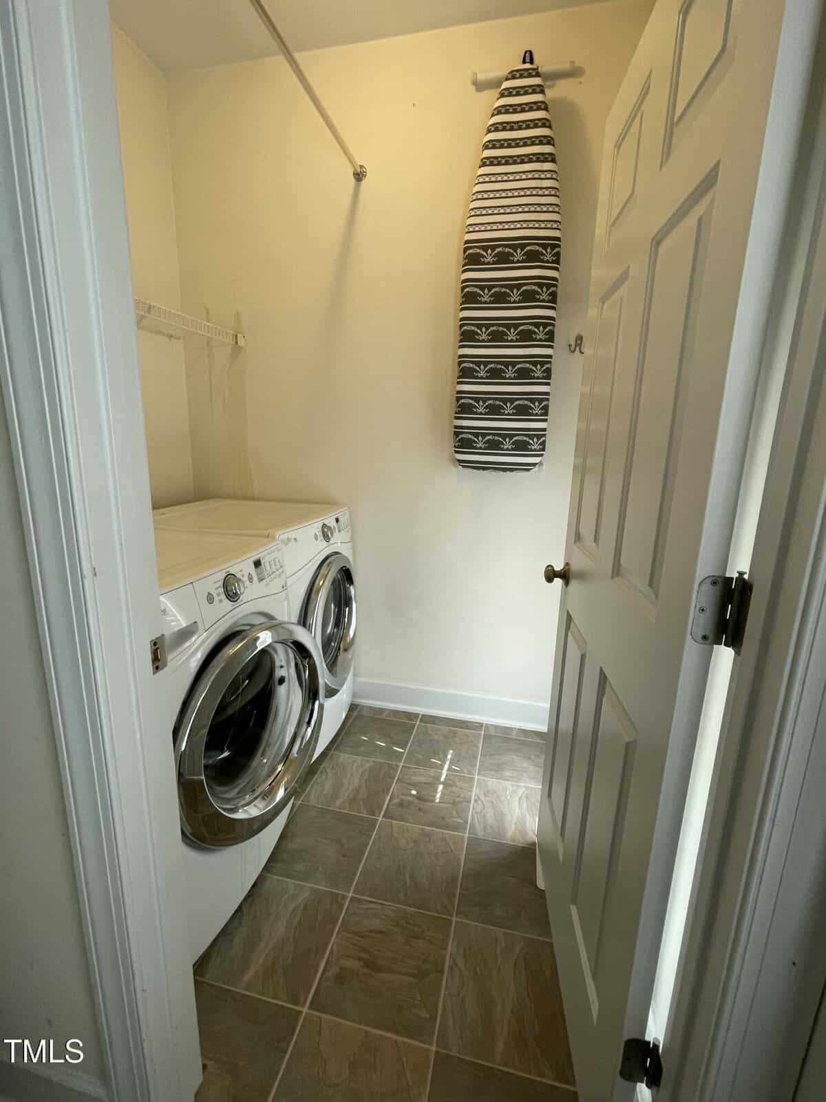 A small laundry room with a front-loading washer and dryer side by side, a wall-mounted ironing board, tiled floor, and a white shelf above the appliances.