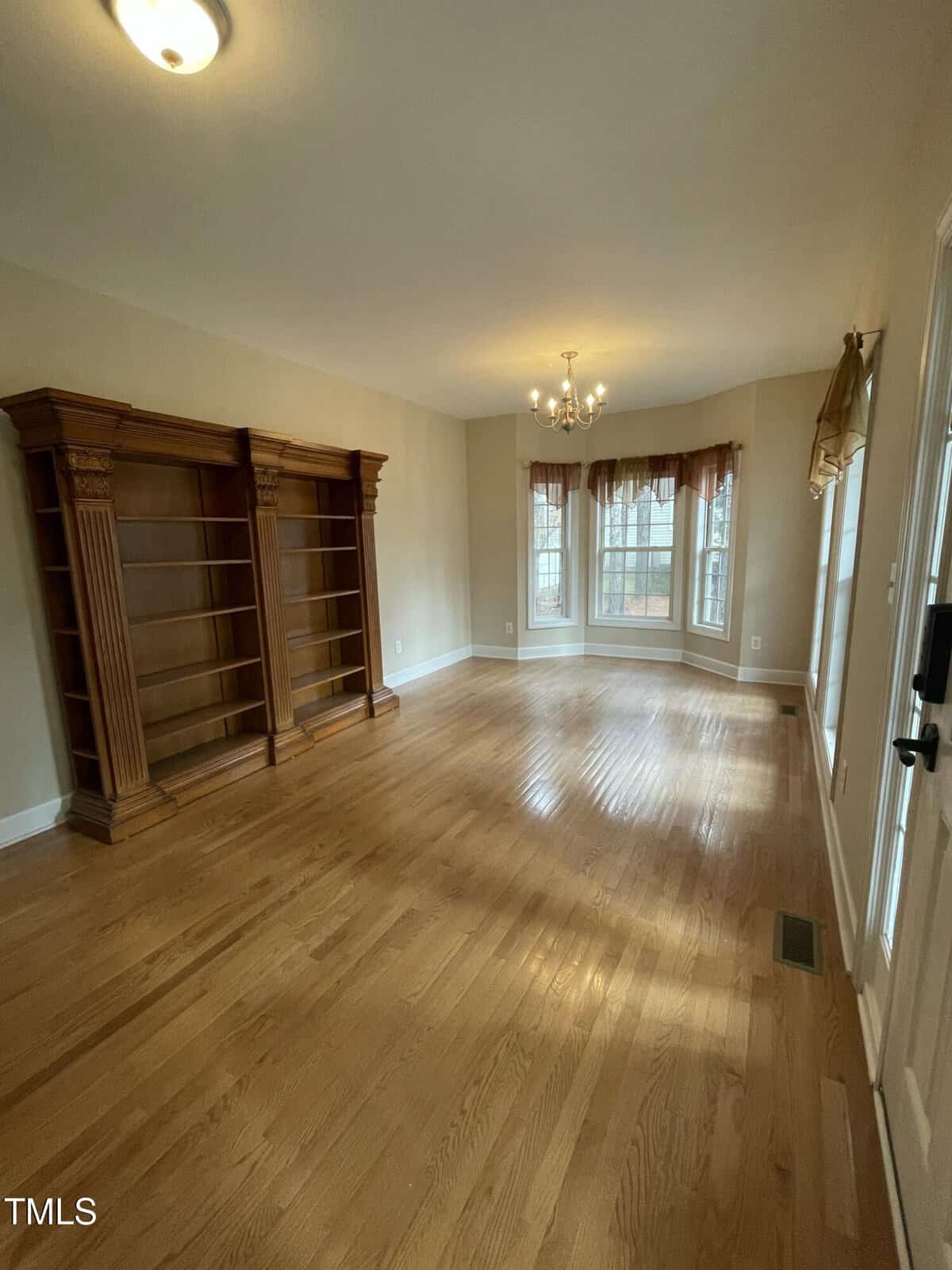 A bright, empty room with hardwood floors, two large wooden bookshelves on the left wall, a hanging chandelier, and a bay window with brown curtains allowing natural light to enter.