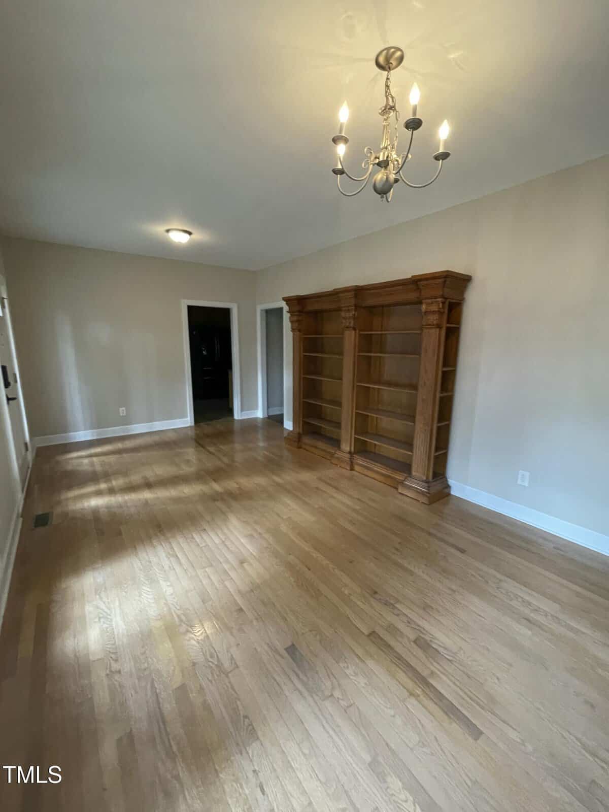 A spacious, empty room with light wood flooring, beige walls, a chandelier, ceiling light, and a large wooden bookshelf along one wall. There are doorways leading to other rooms.