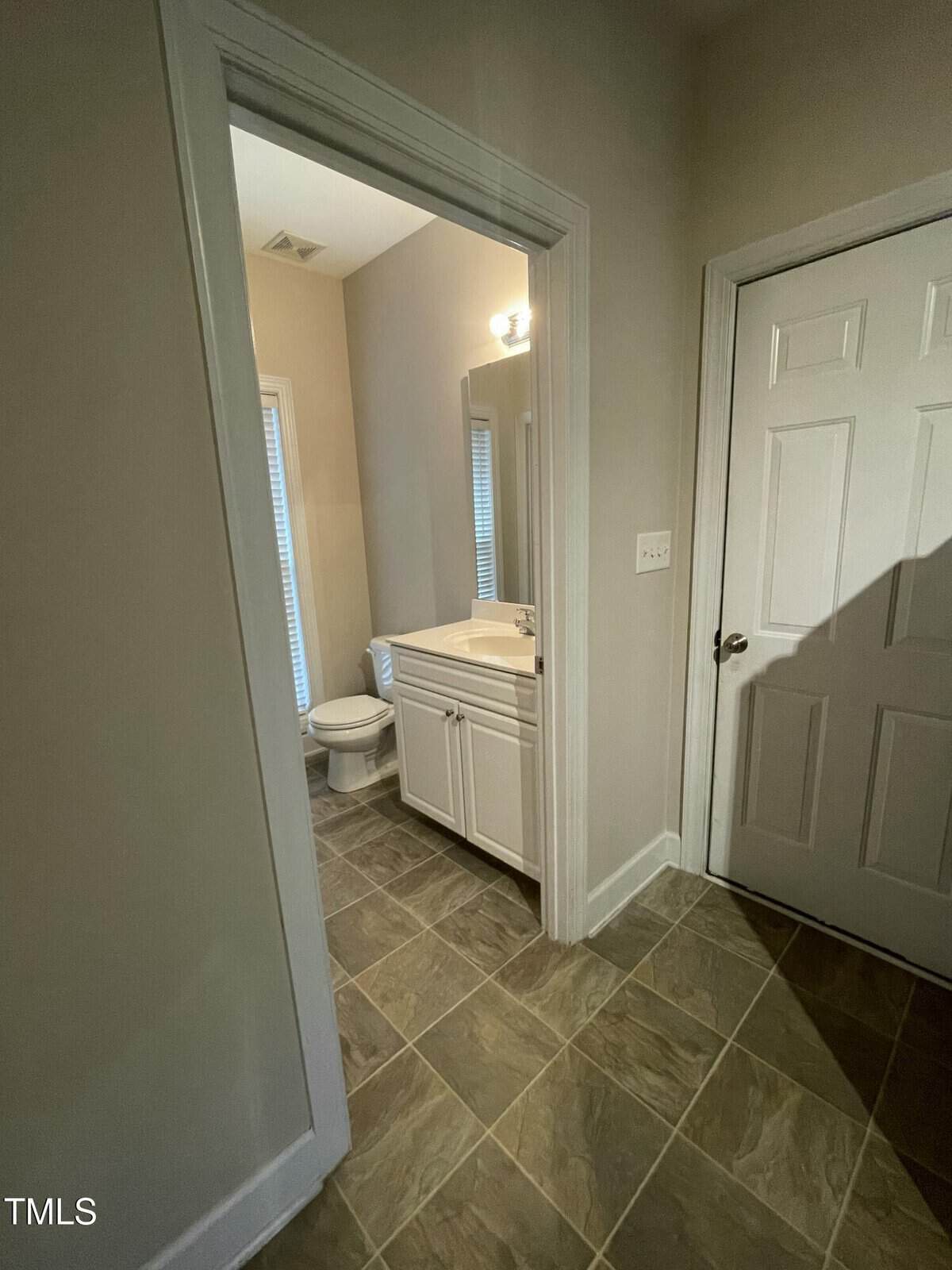 A small bathroom with beige walls and tile flooring, featuring a white vanity with a sink, a mirror above, and a toilet next to a window with blinds. A white door is visible in the hallway.