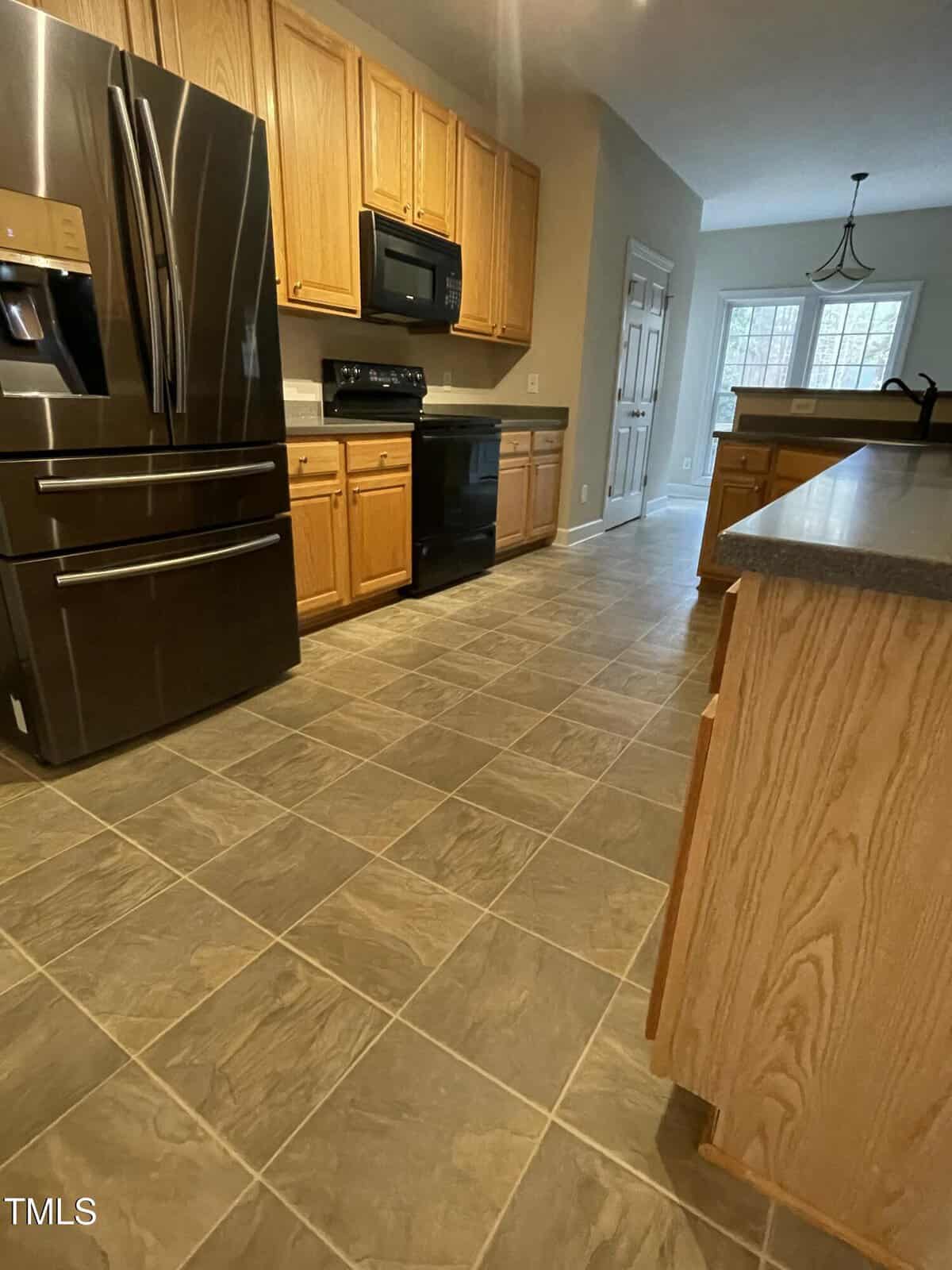 A kitchen with wooden cabinets, dark countertops, tile flooring, stainless steel refrigerator, black stove and microwave, and a windowed dining area in the background.