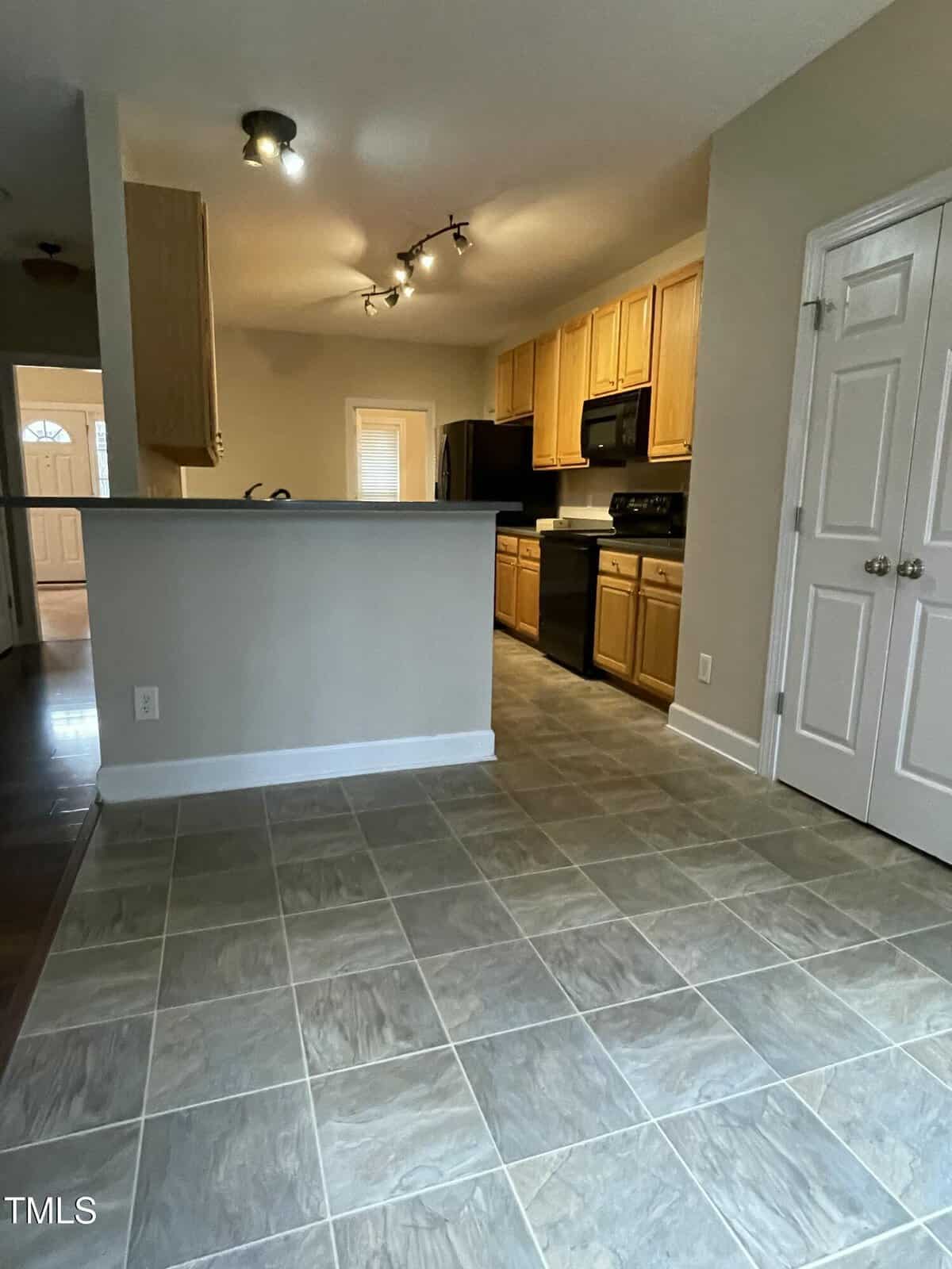 A kitchen with light wood cabinets, black appliances, beige walls, tile flooring, and track lighting. A half wall separates the kitchen from the adjacent tiled dining area. Double doors are visible on the right.